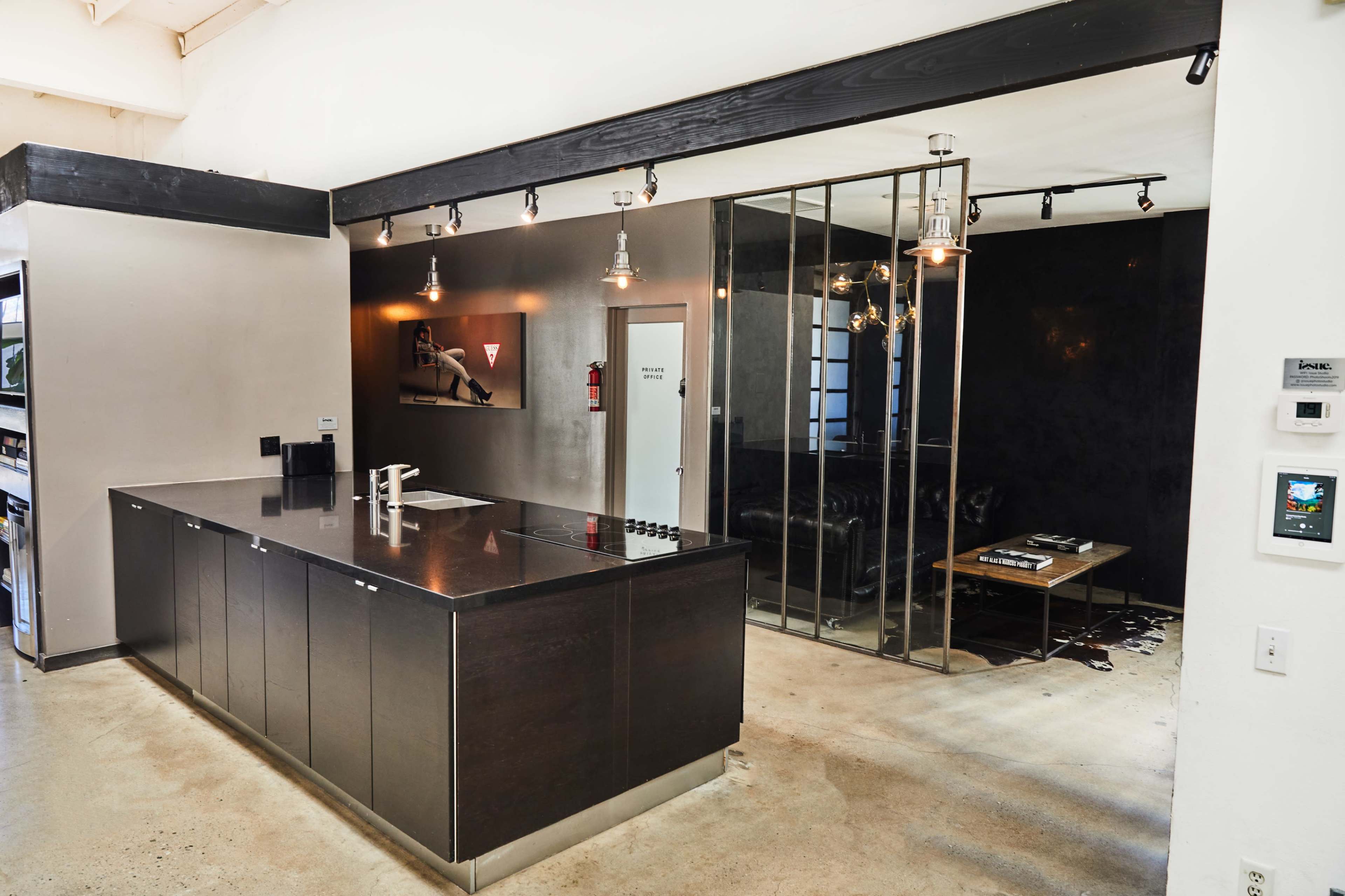 The image shows a modern kitchen with a dark countertop and an adjacent glass-walled room that includes a black leather couch and a wooden table.