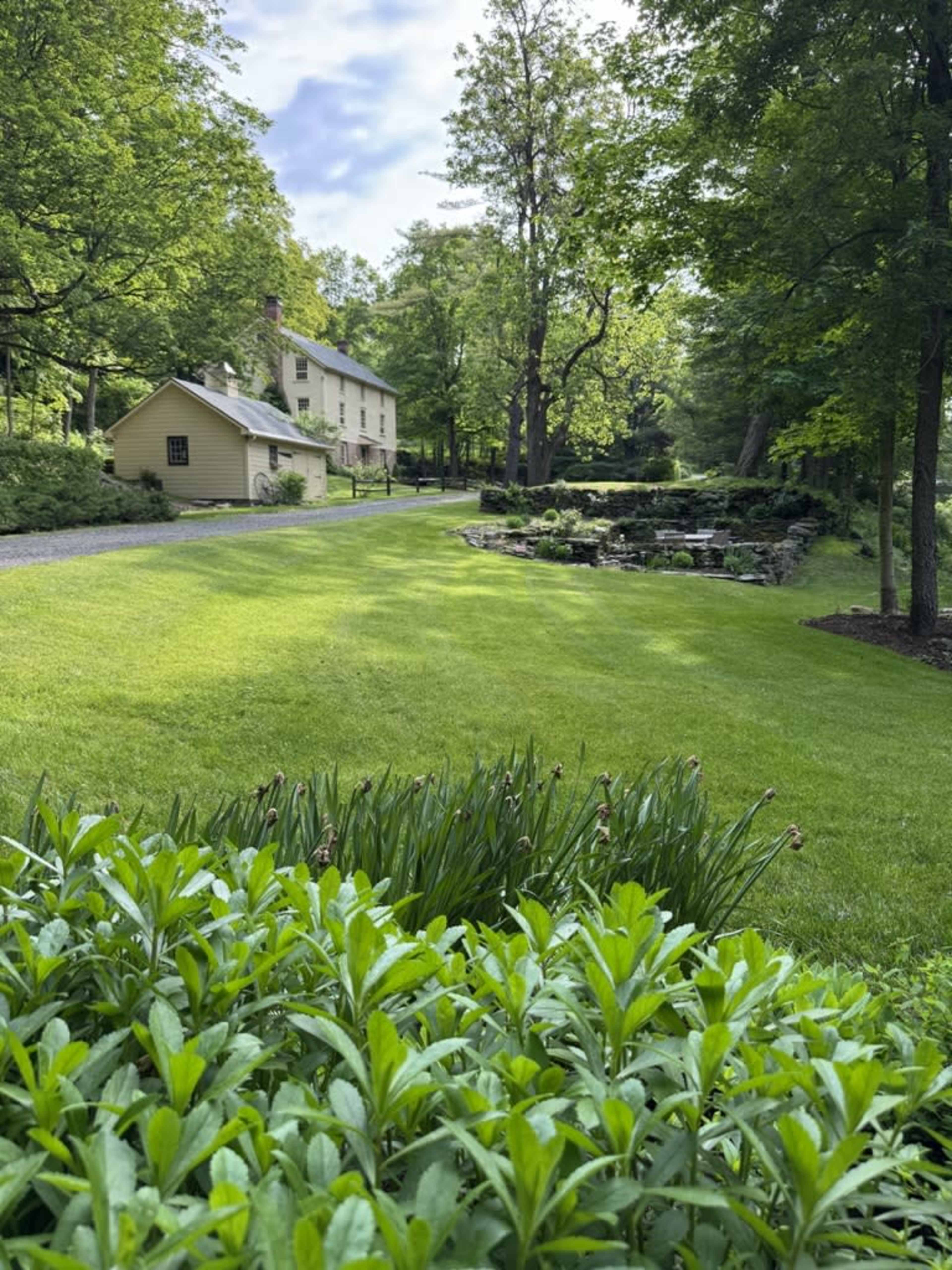A well-maintained lawn leads to a two-story house surrounded by trees and a small water feature in the background.