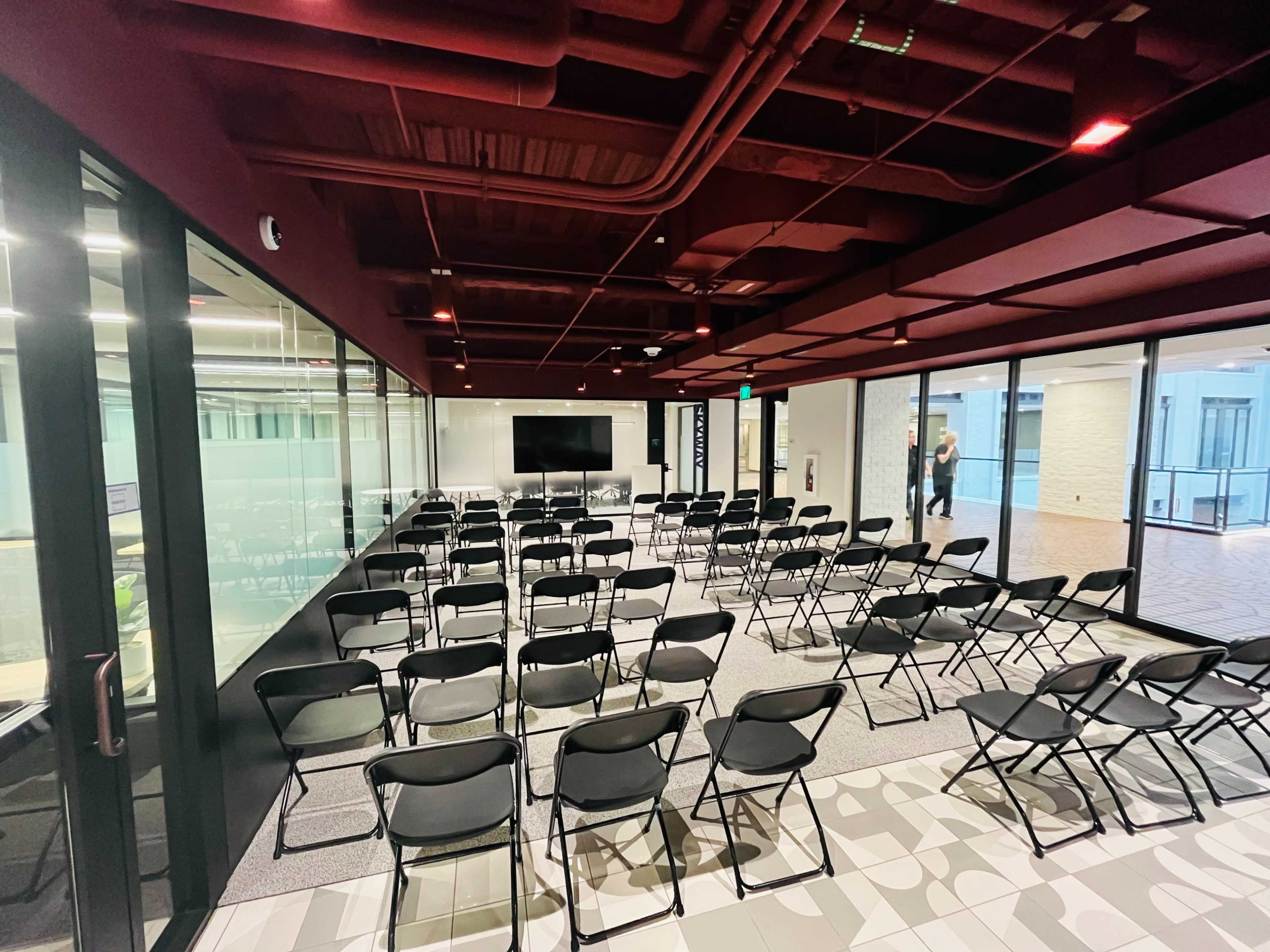 A meeting room set up with rows of black folding chairs facing a screen and glass walls.
