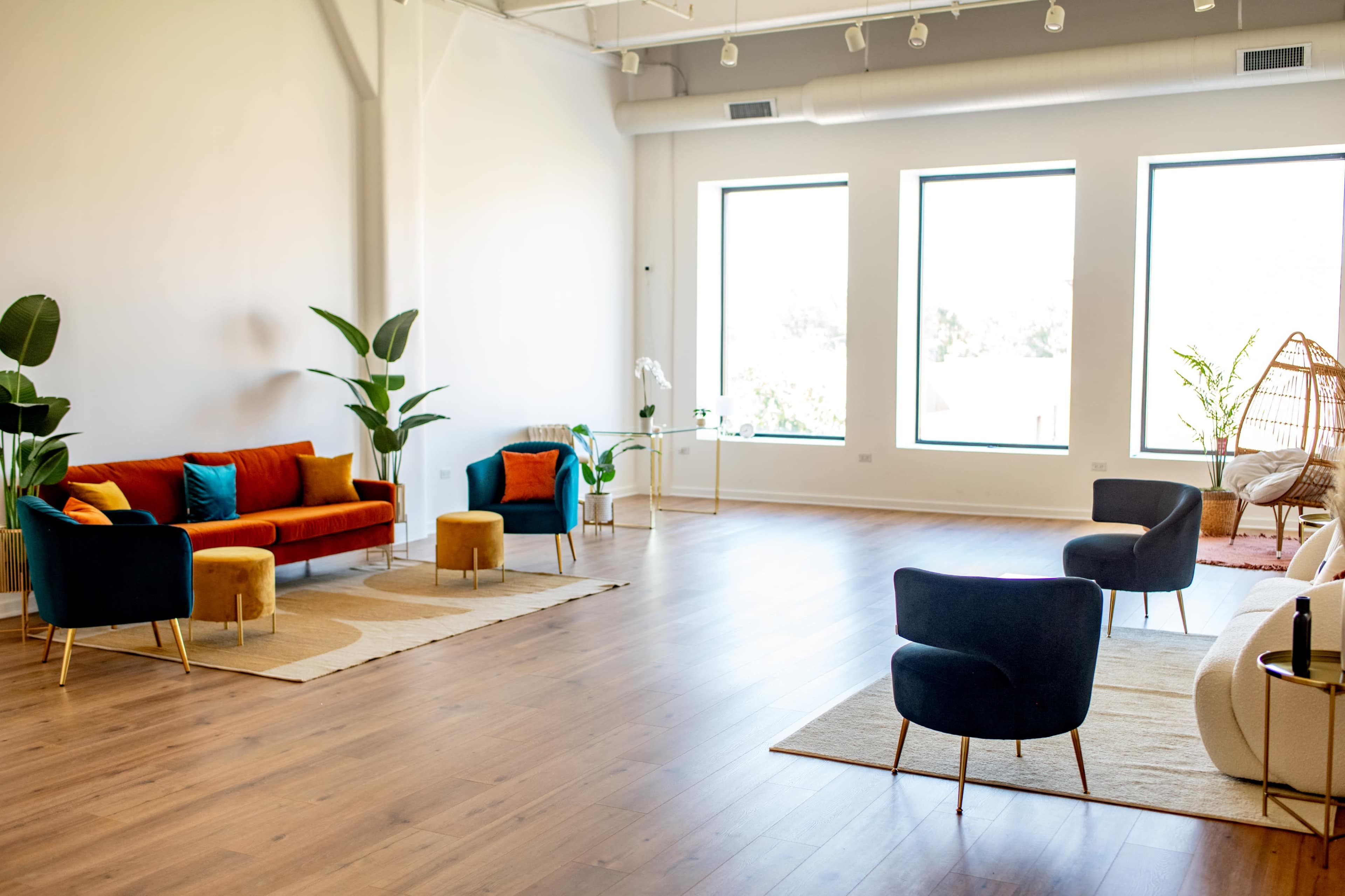 A spacious room features modern furniture with colorful couches and chairs, surrounded by large windows and indoor plants.