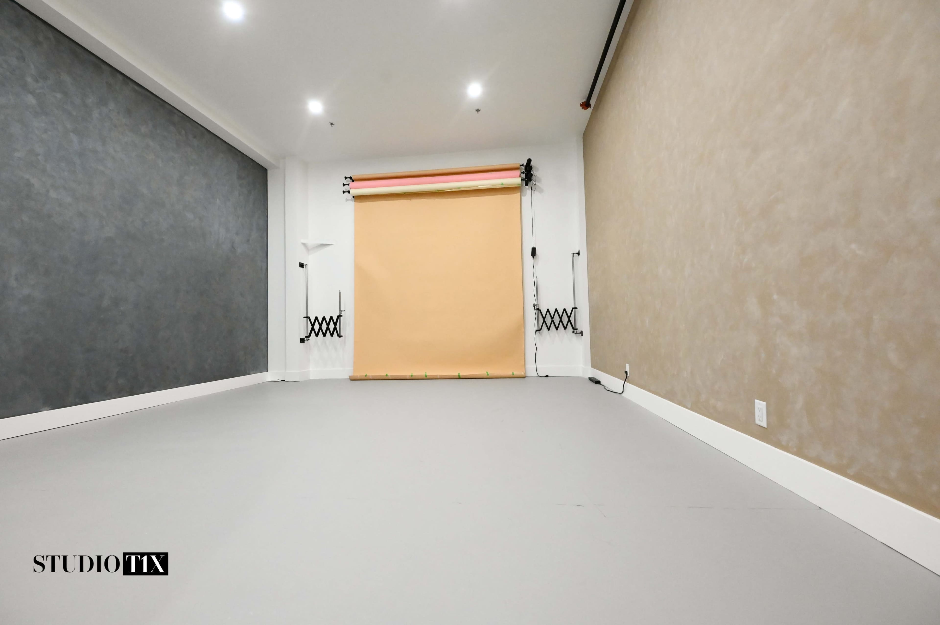 The image shows a minimalist photography studio with a gray wall, a tan backdrop on a roll, and a smooth light-colored floor.