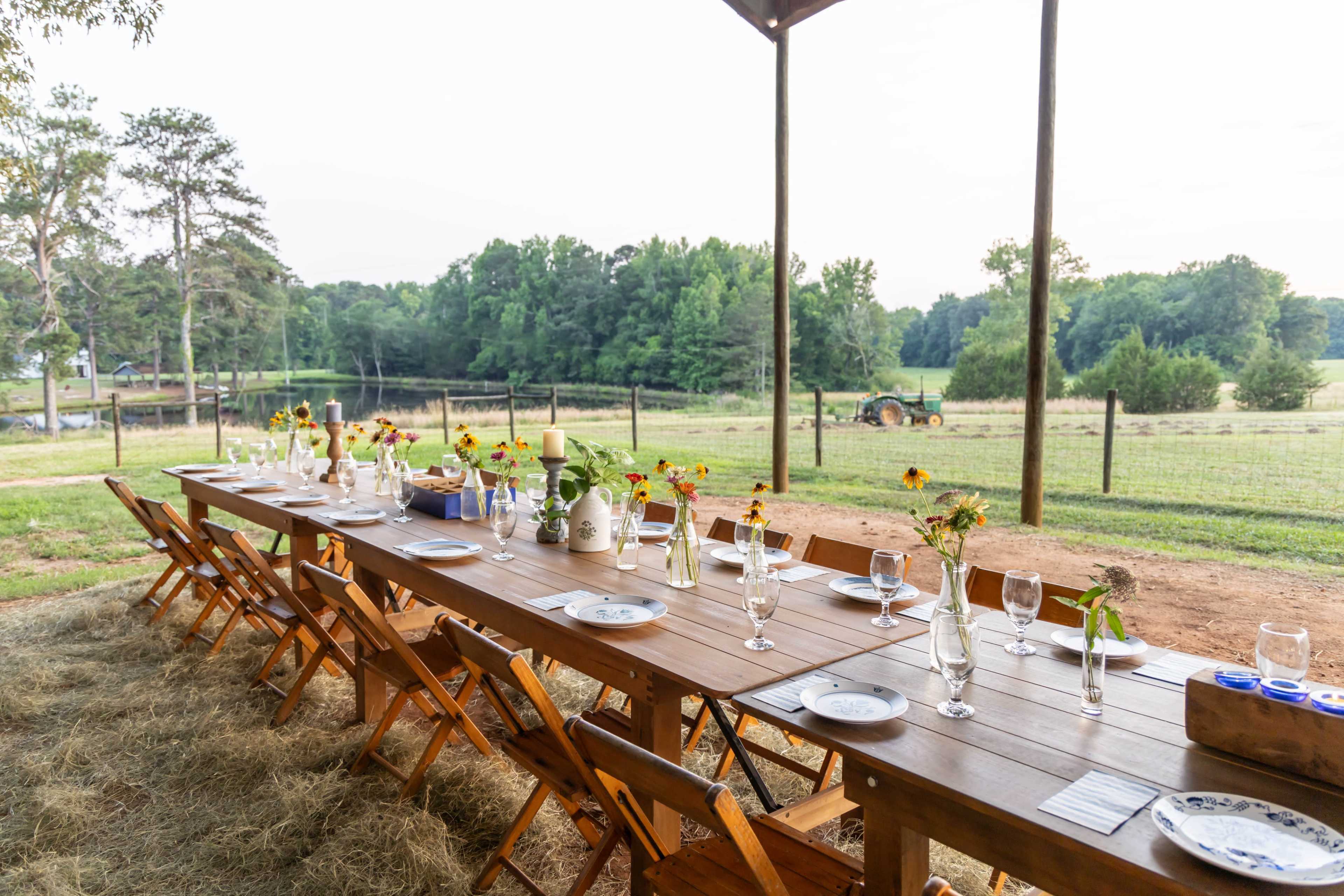 A long wooden table is set for a meal outdoors, adorned with vases of sunflowers, overlooking a grassy field and a distant tractor.