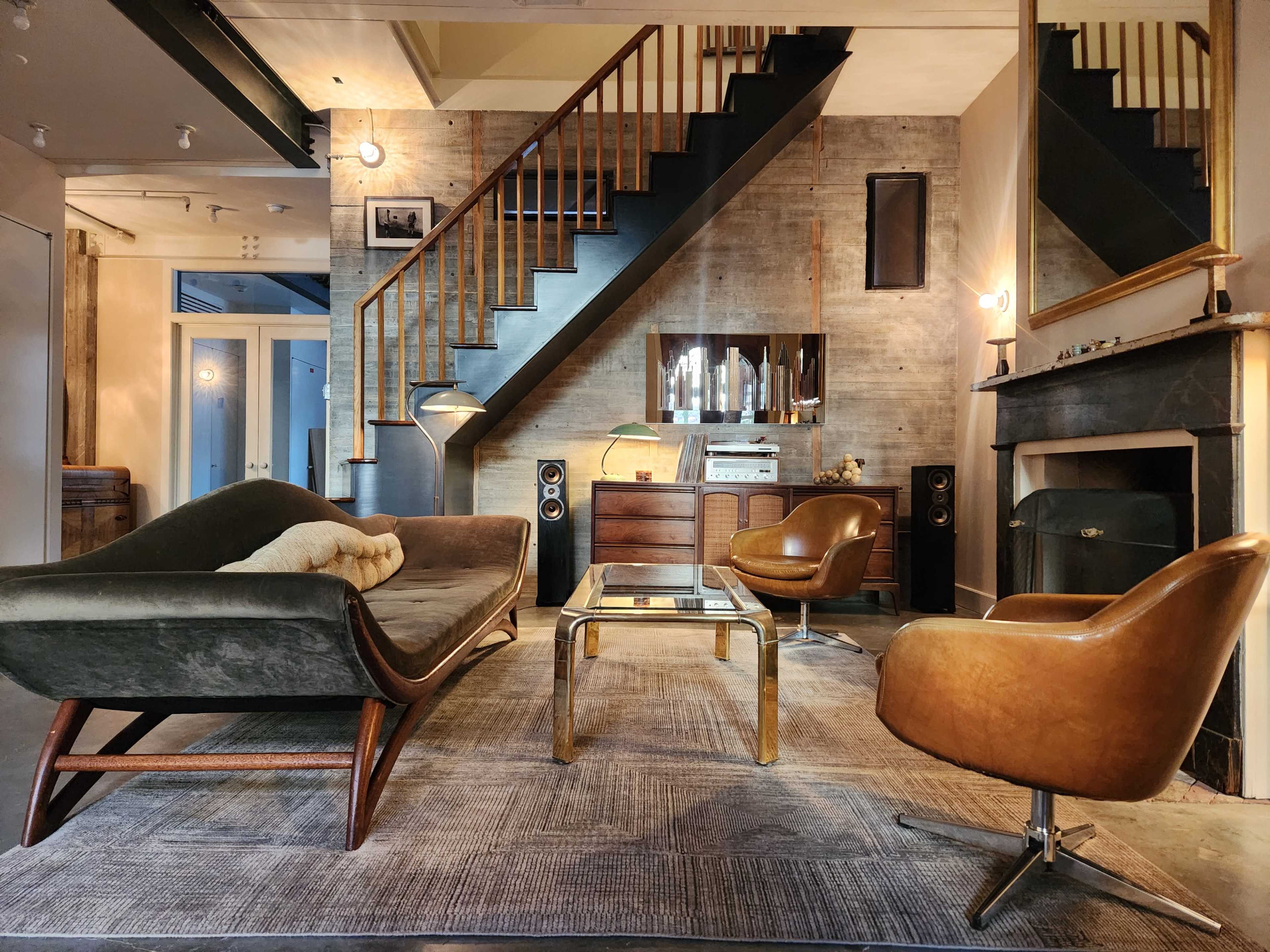 The image shows a mid-century modern living room with a staircase, featuring a curved sofa, two stylish chairs, a glass coffee table, and a record player in wooden cabinetry.