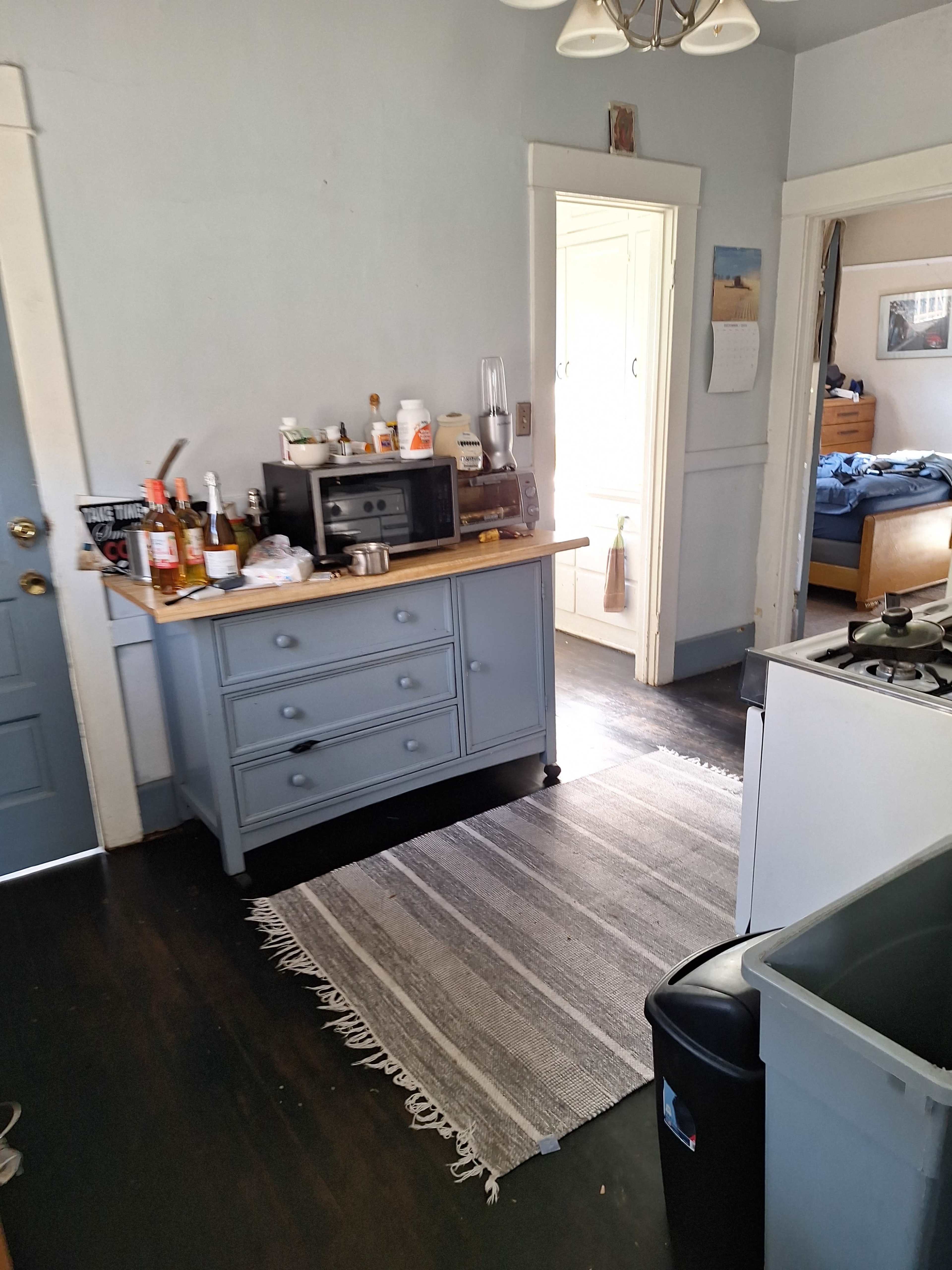 The image shows a small kitchen area featuring a blue cabinet, a microwave, and a rug, with a doorway leading to a bathroom and another room in the background.