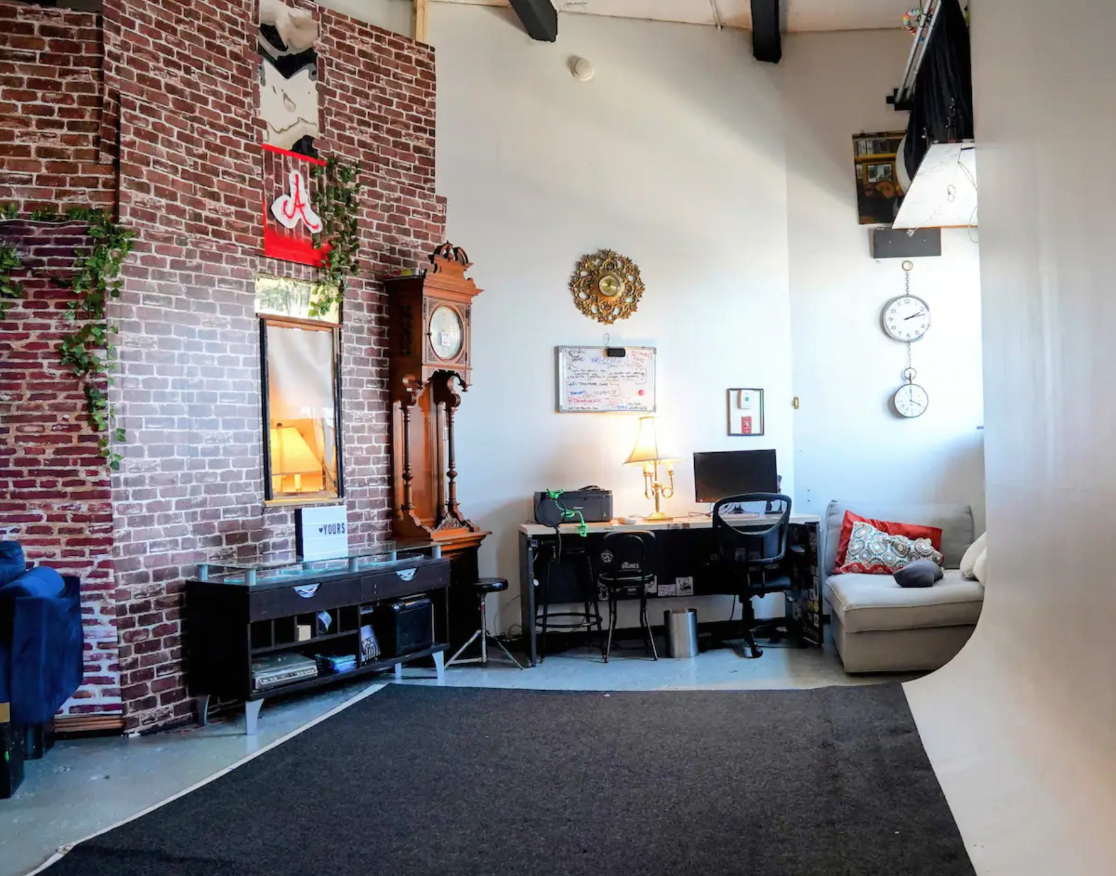 The scene depicts a spacious room featuring a brick wall, a clock, a desk with a computer, a mirror, and a cozy seating area.