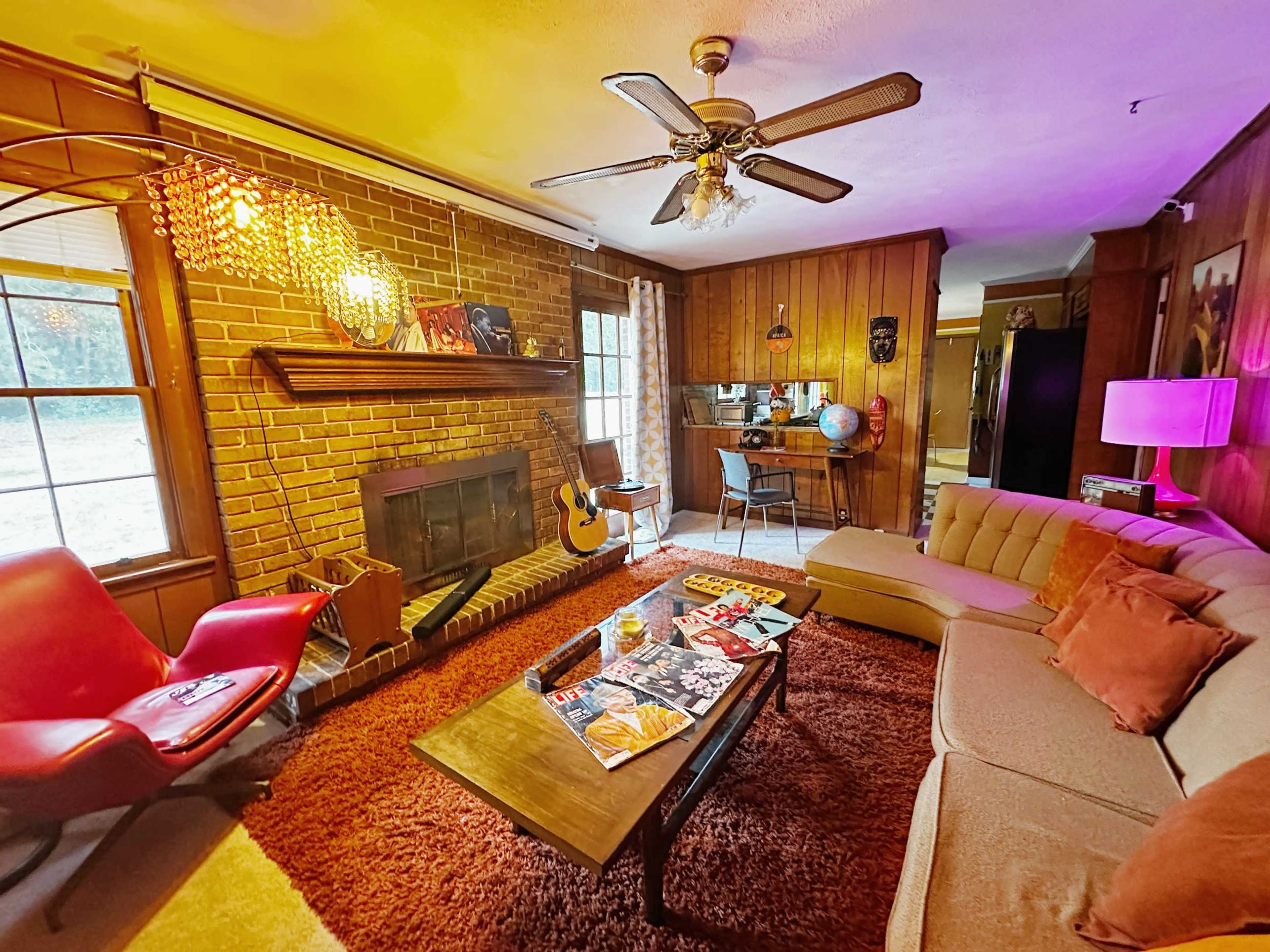 The living room features a brick fireplace, a sectional sofa, a coffee table with magazines, and large windows allowing natural light to enter.