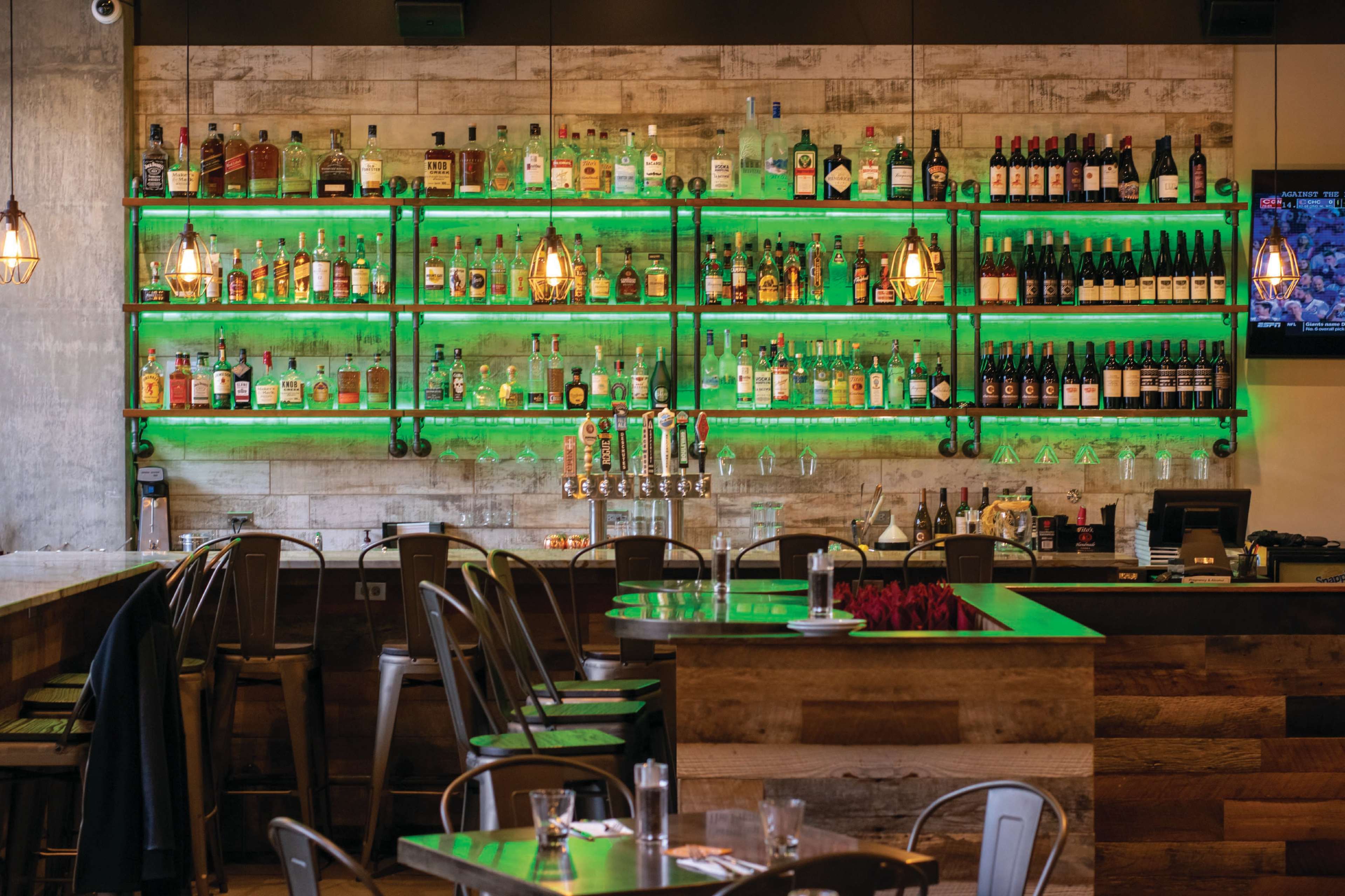 The image shows a restaurant interior featuring a bar with illuminated shelves displaying various bottles of alcohol and several seating areas.