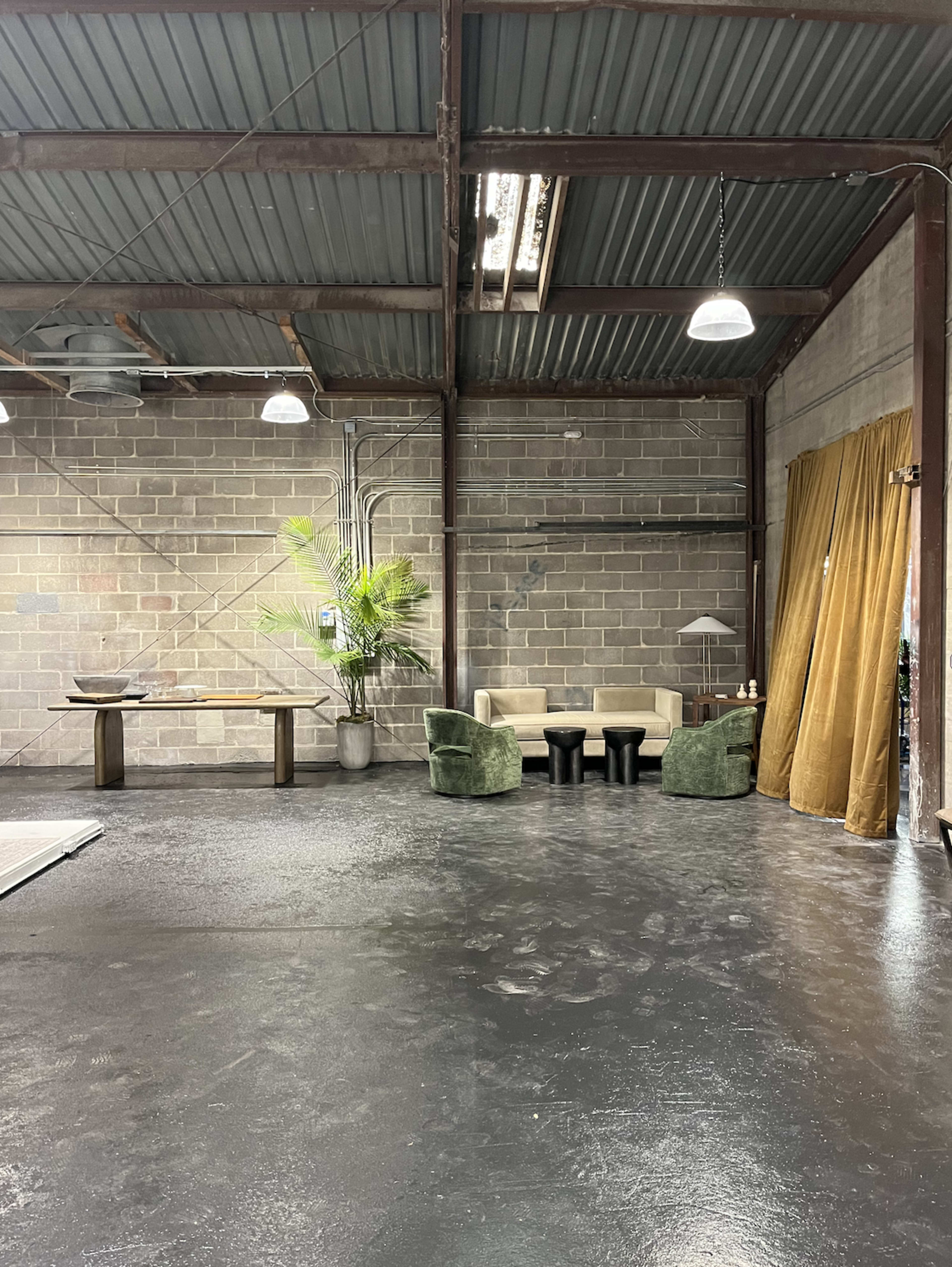 An industrial-style interior with concrete walls, a dark floor, a small seating area with two green chairs and a beige sofa, a potted plant, and a curtain draped in the corner.