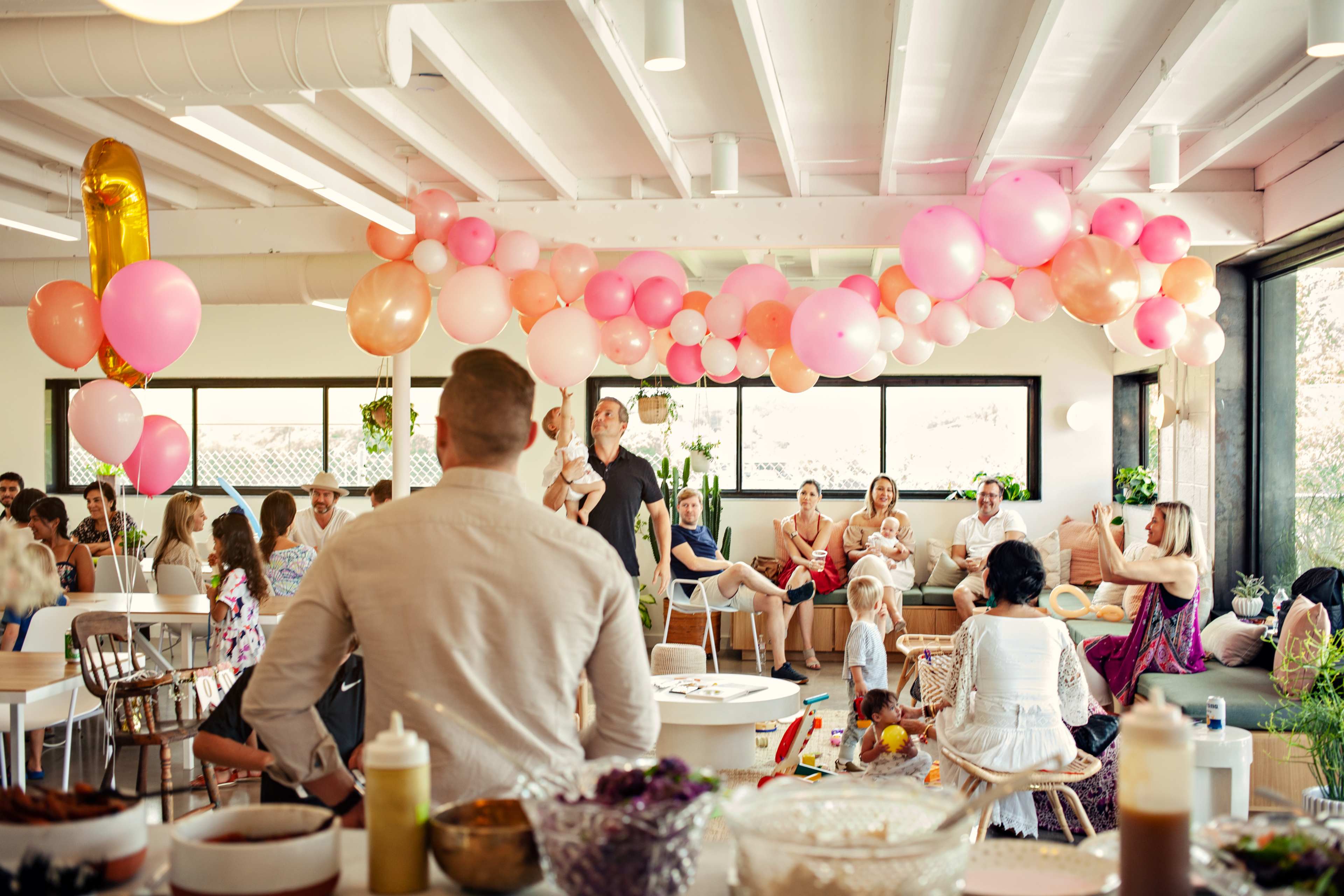 A lively gathering is taking place in a bright room decorated with pink and gold balloons, where guests sit at tables and socialize.
