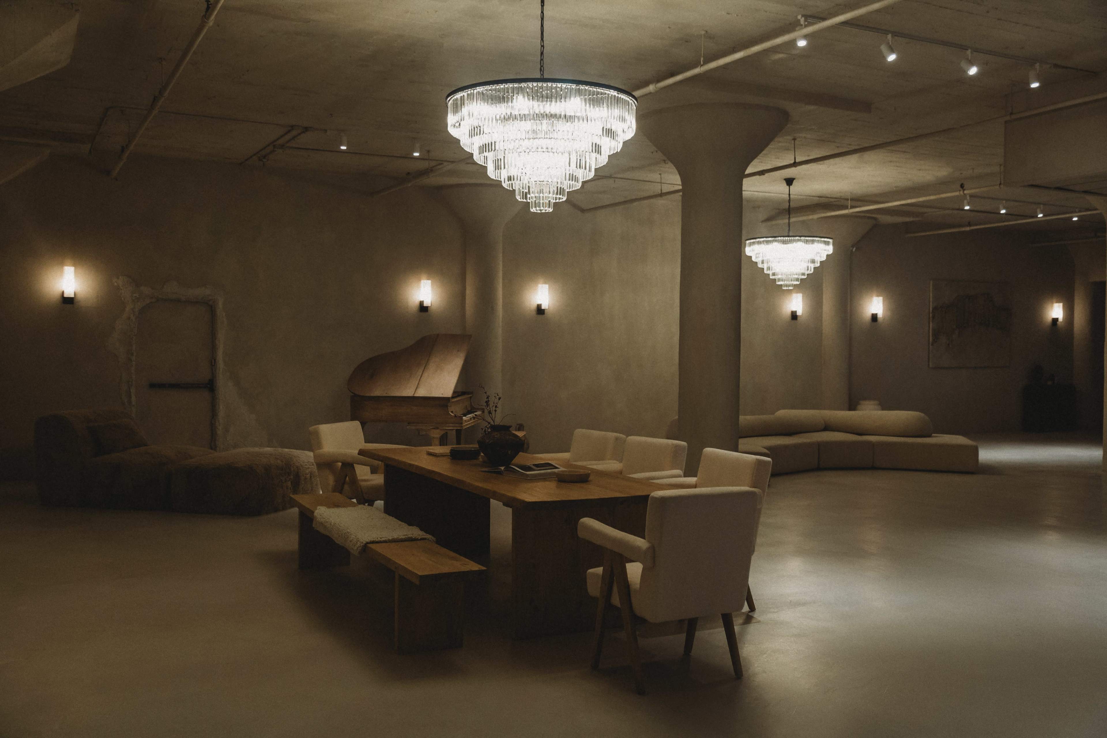 A spacious, dimly lit room with a large wooden dining table surrounded by white chairs, a grand piano in the corner, and two distinct chandeliers overhead.