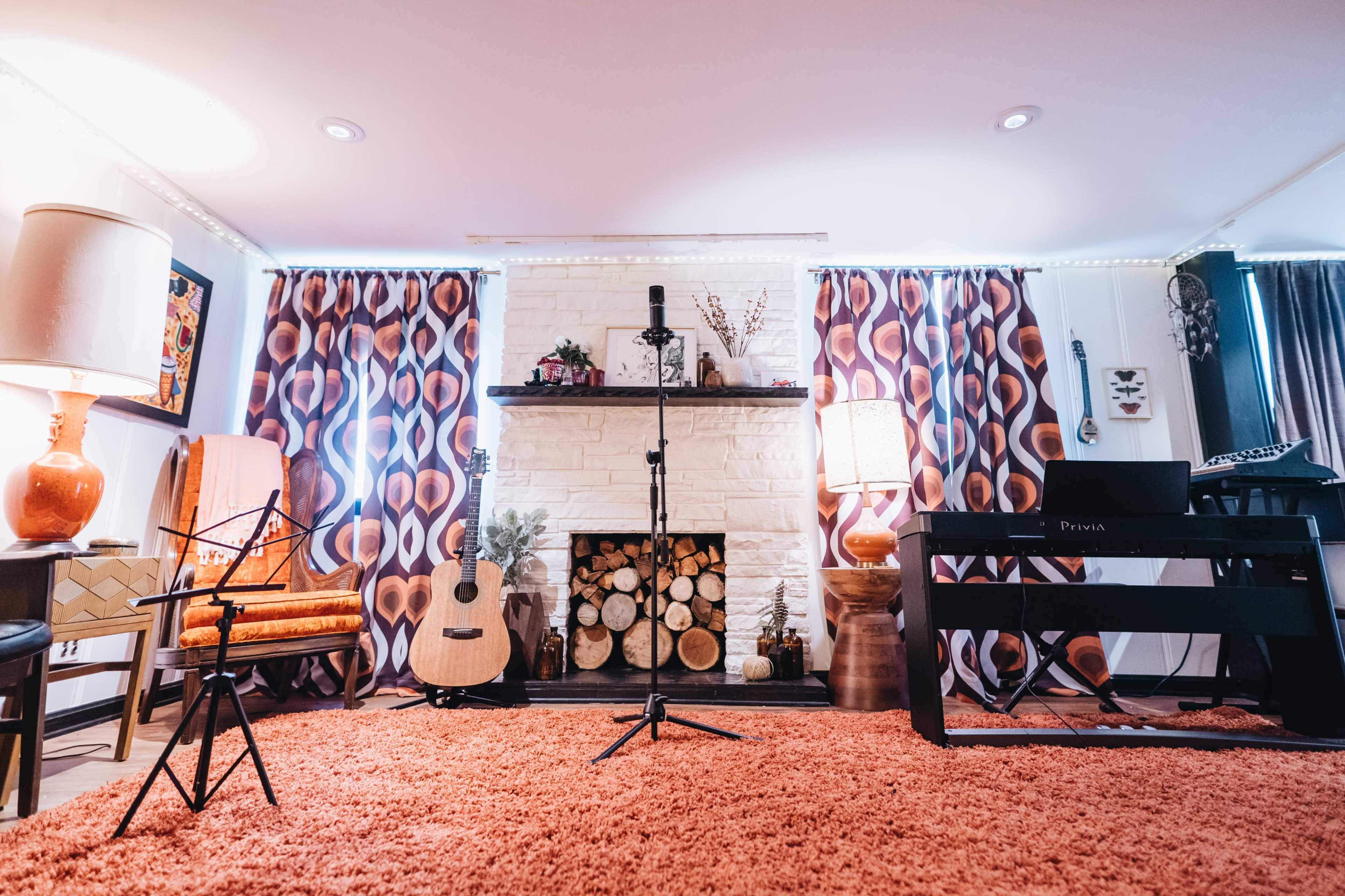 A cozy living room with a fireplace, decorated with guitars, a microphone on a stand, and two lamps, all set on an orange rug.
