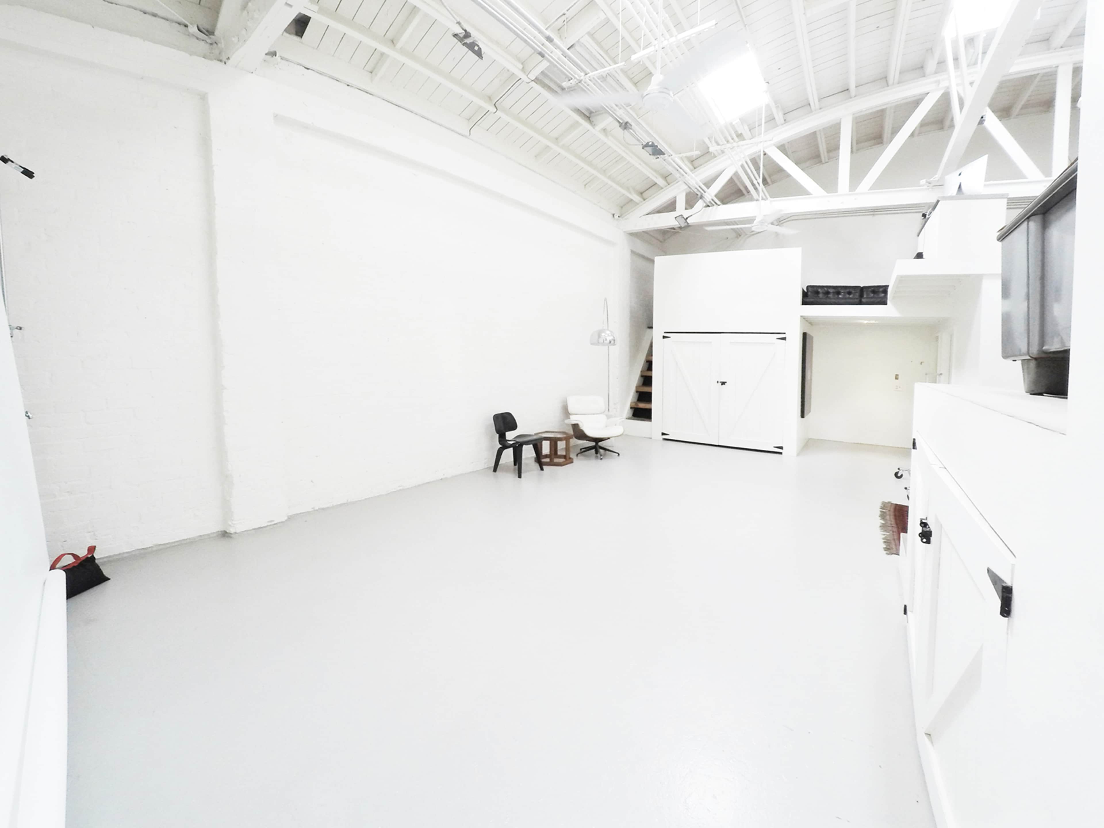 A spacious, minimally furnished studio with white walls and a light gray floor, featuring a chair, a small table, and a wall-mounted cabinet.