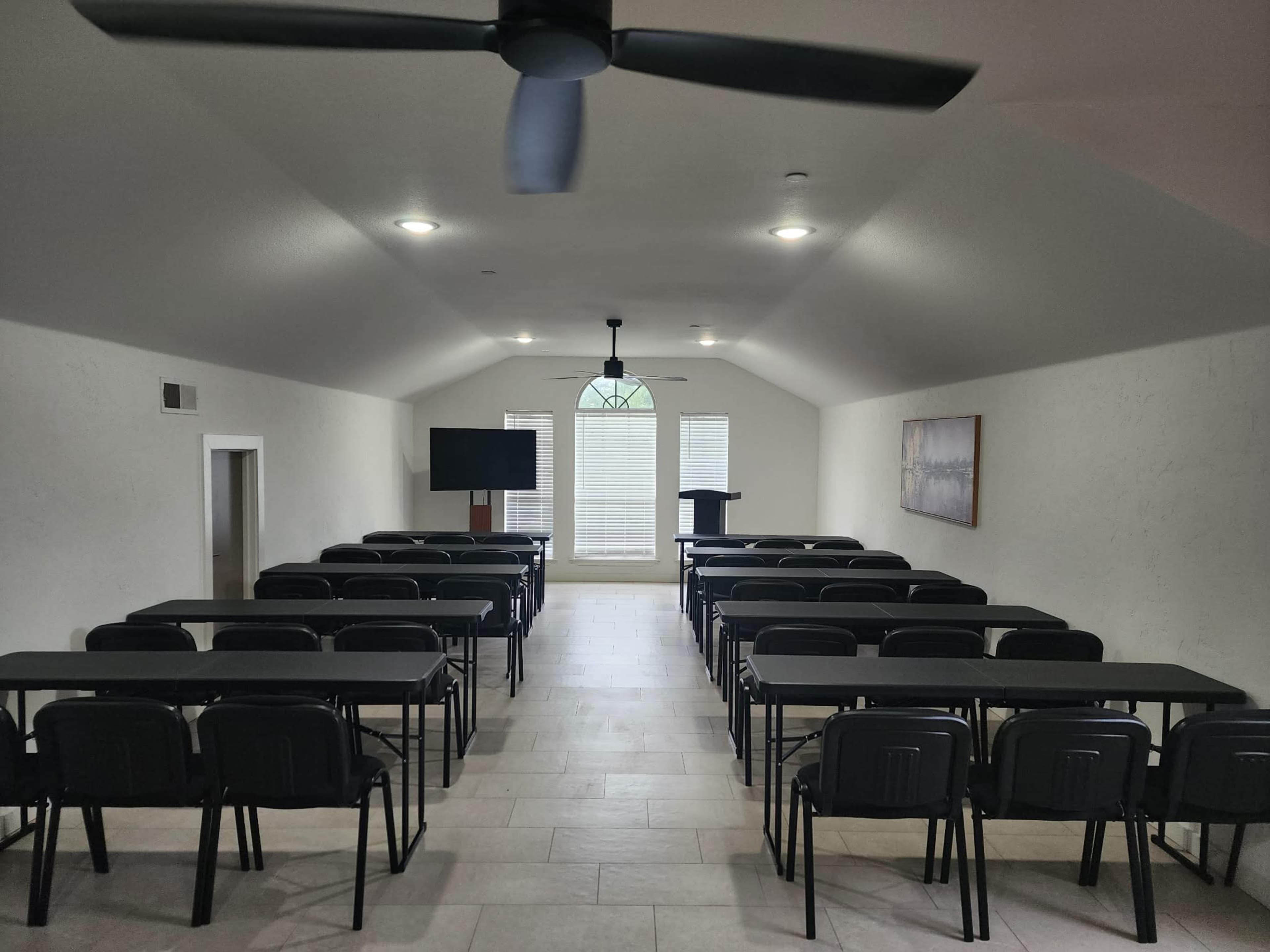 The room features rows of black folding chairs arranged facing a podium, with a large screen and windows at the back.