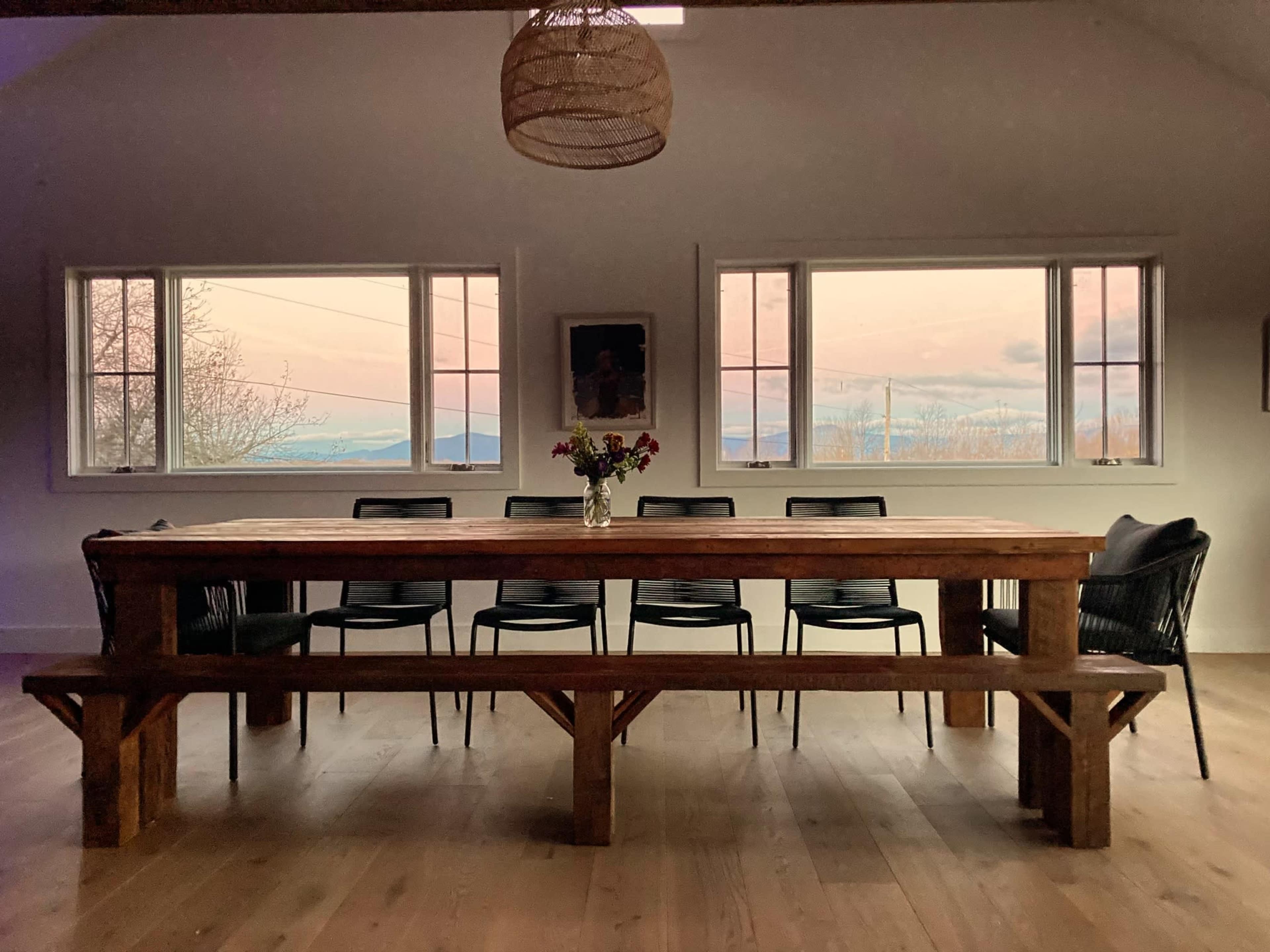 A long wooden dining table with black chairs sits in a bright room, framed by large windows offering a view of a sunset landscape.