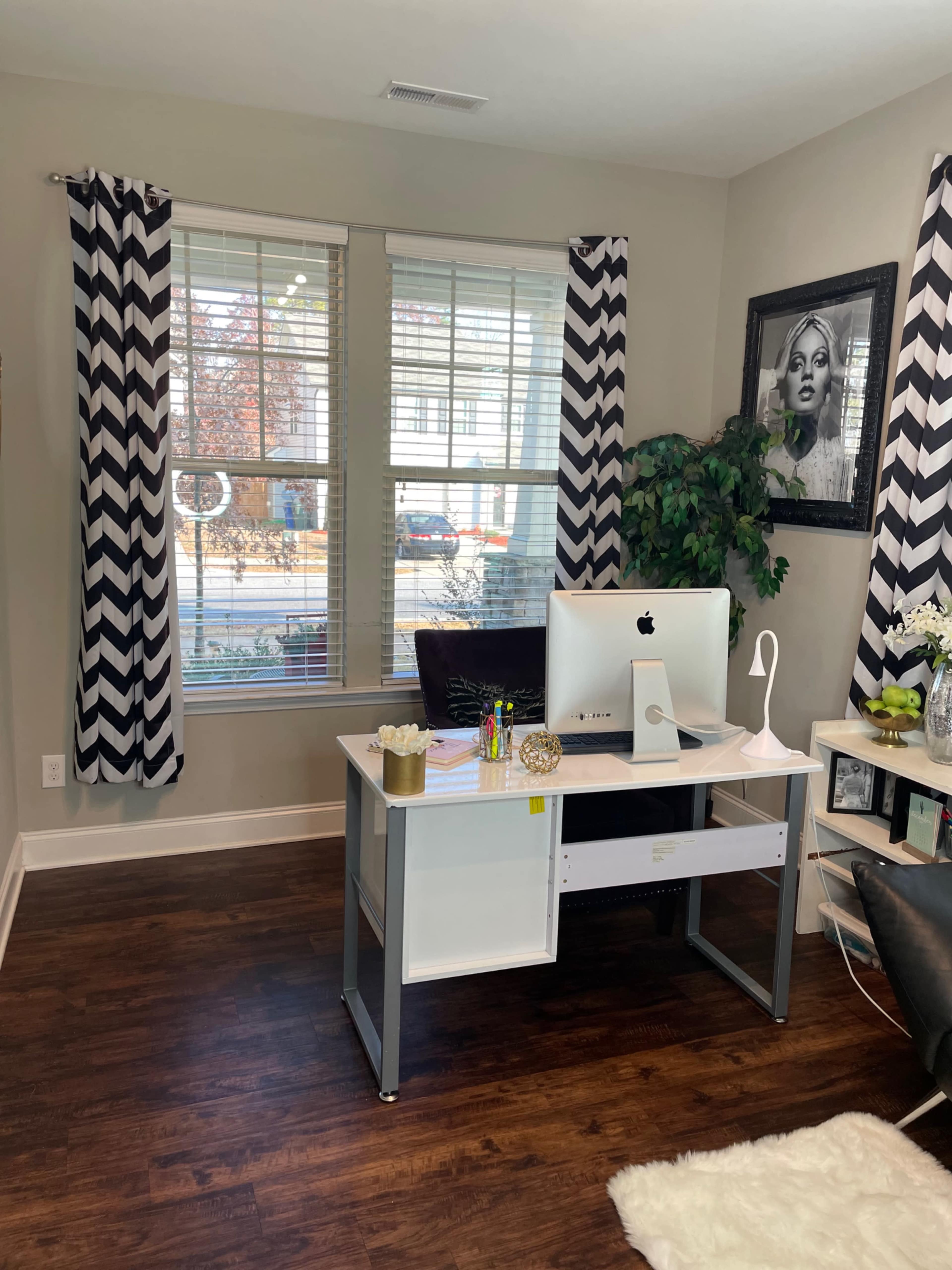 A home office features a desk with a computer, colorful pens, and decorative items, set against large windows with chevron-patterned curtains.