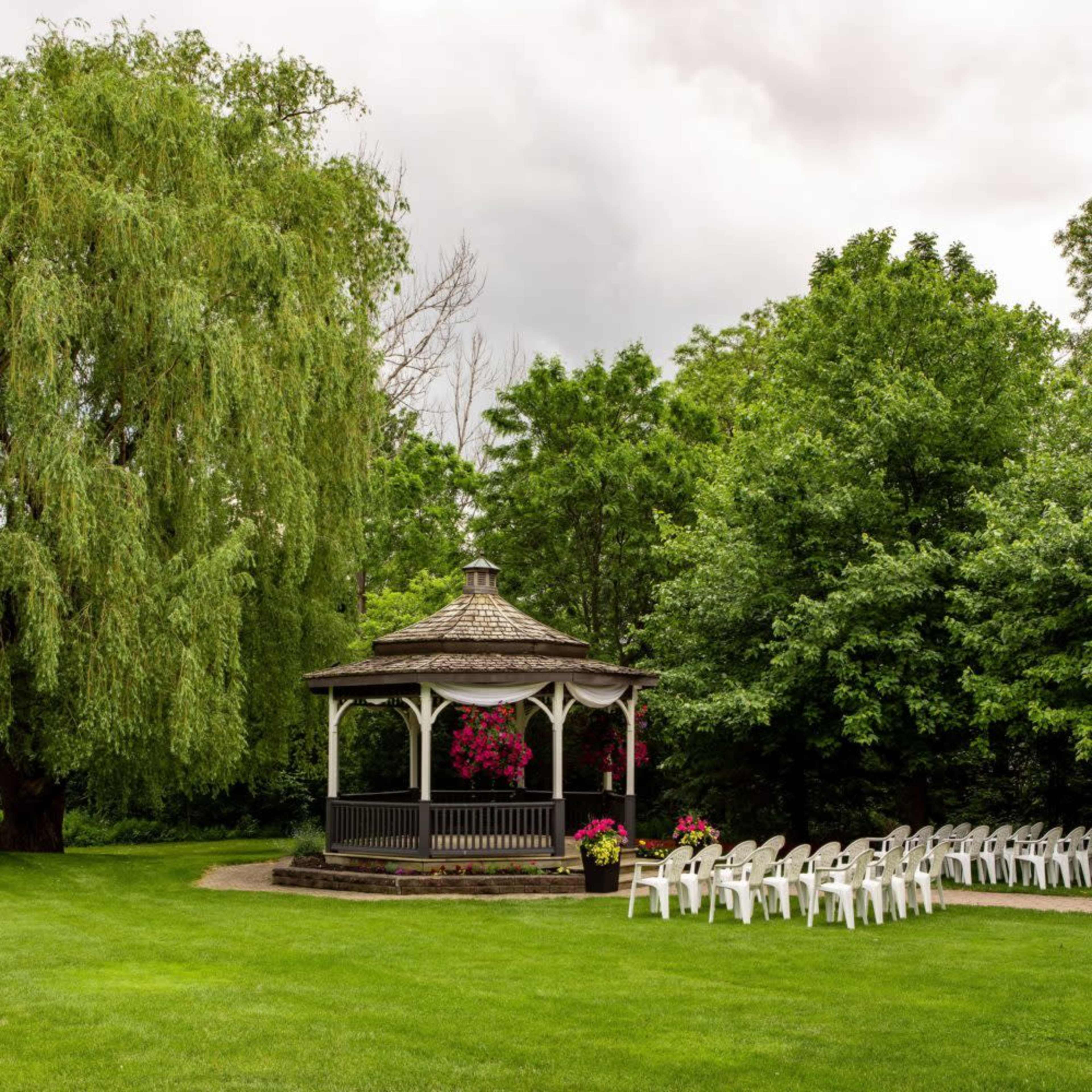 A gazebo surrounded by trees and a lawn features white chairs arranged in front of it.