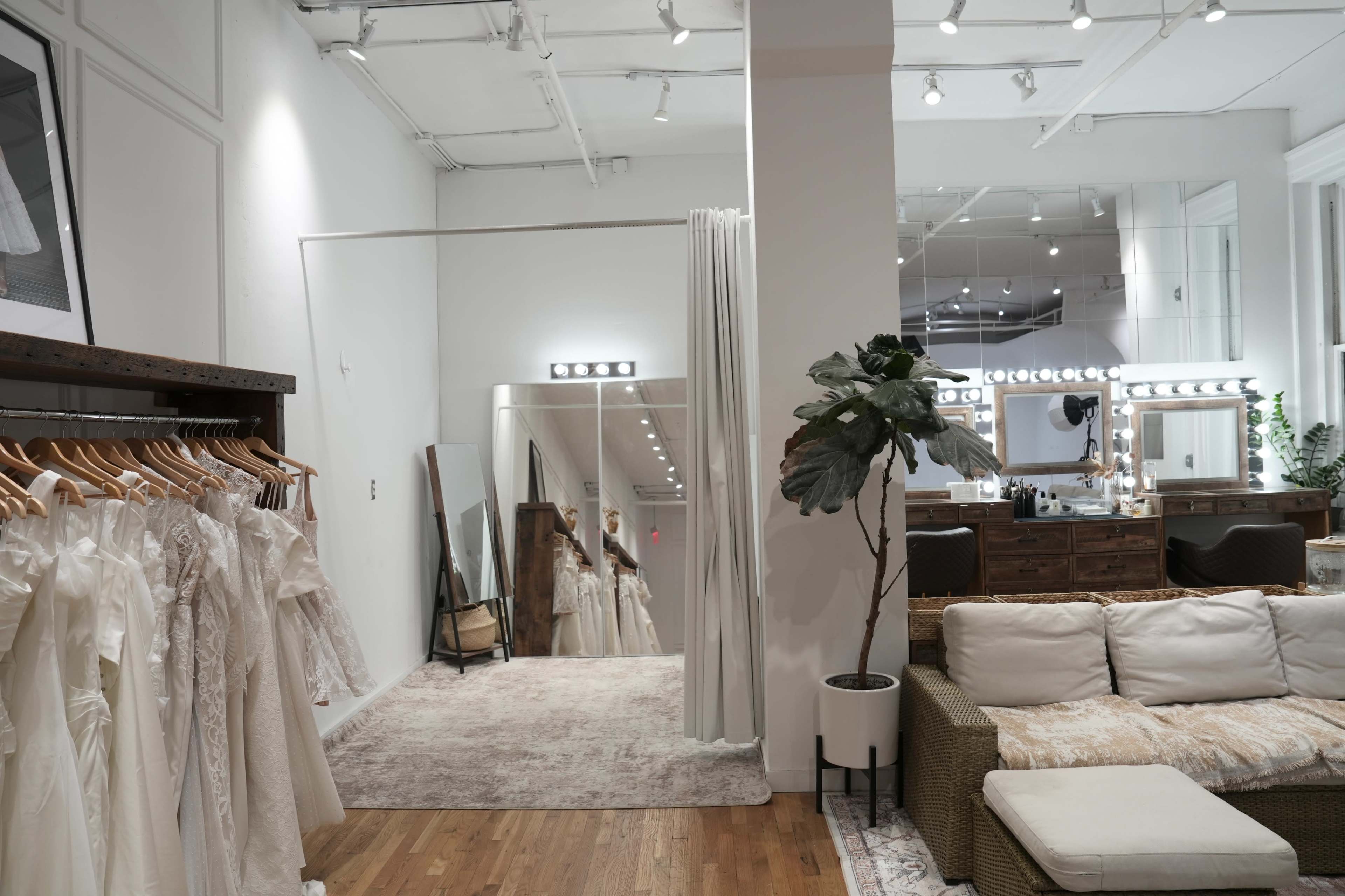 The image depicts a bridal shop interior featuring wedding dresses on display, mirrors, and a lounge area with a sofa and plants.