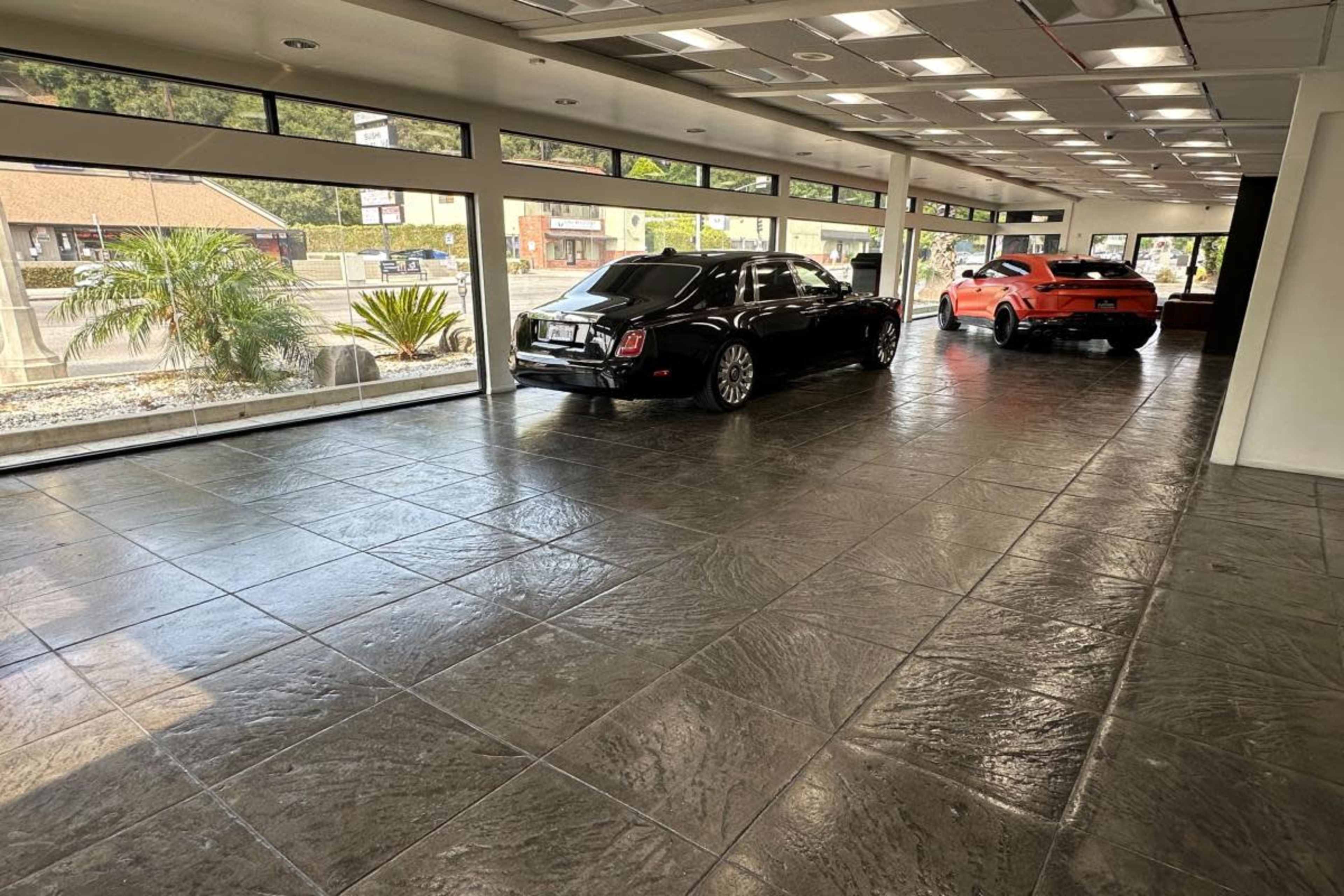 The image shows two luxury cars, a black sedan and an orange sports car, parked inside a spacious, well-lit showroom with large windows.
