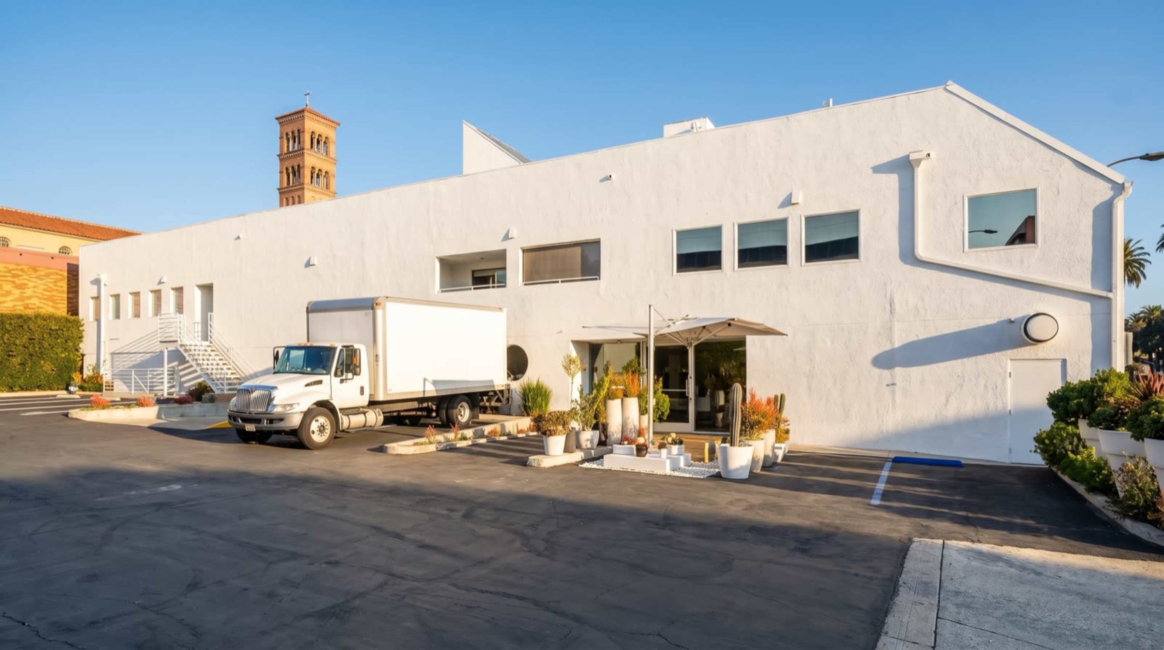 A large white building with several windows and a truck parked in front is set against a clear blue sky.
