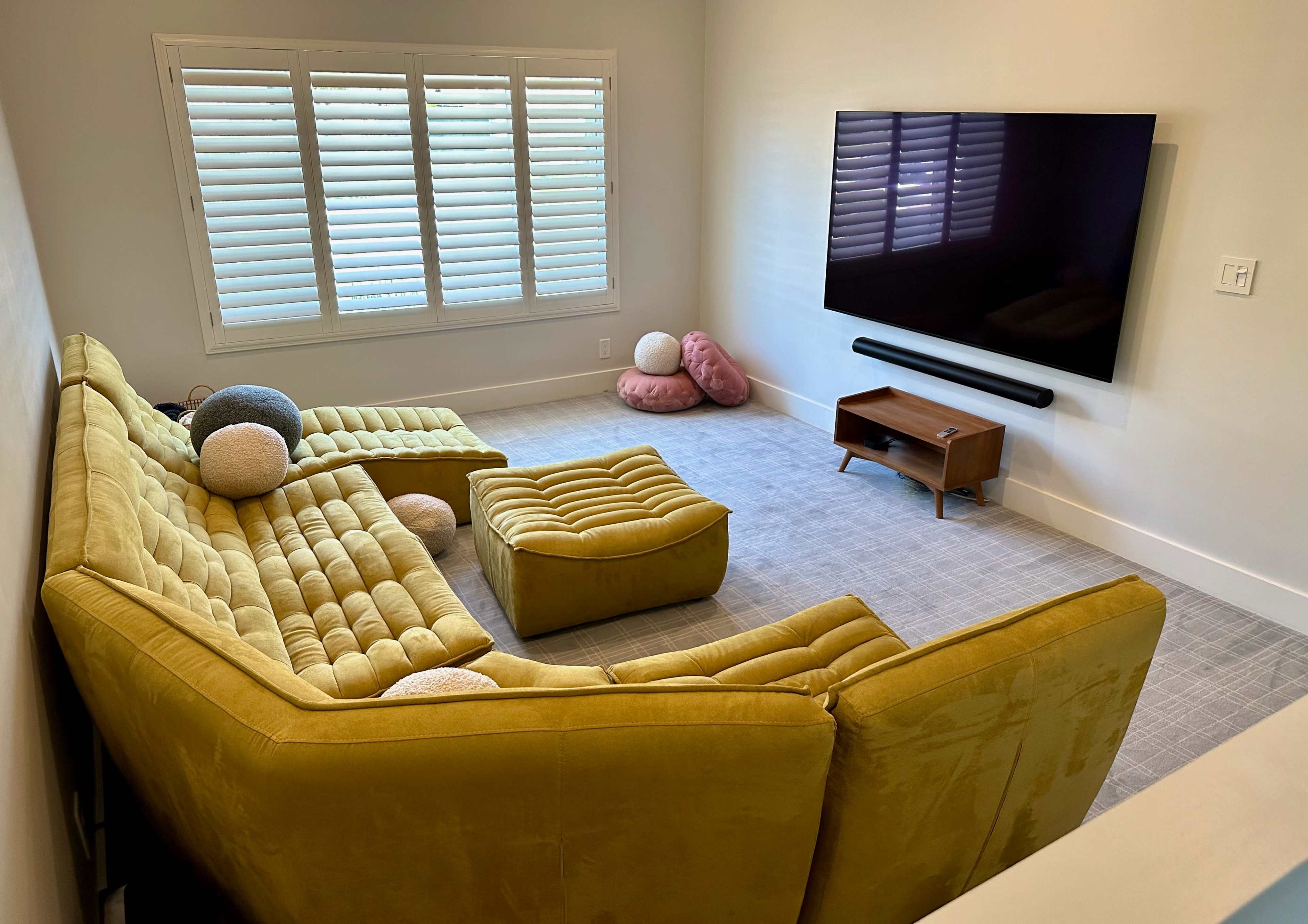 The room features a yellow sectional sofa, a matching ottoman, a wooden TV stand, and several decorative pillows on a light gray carpet.