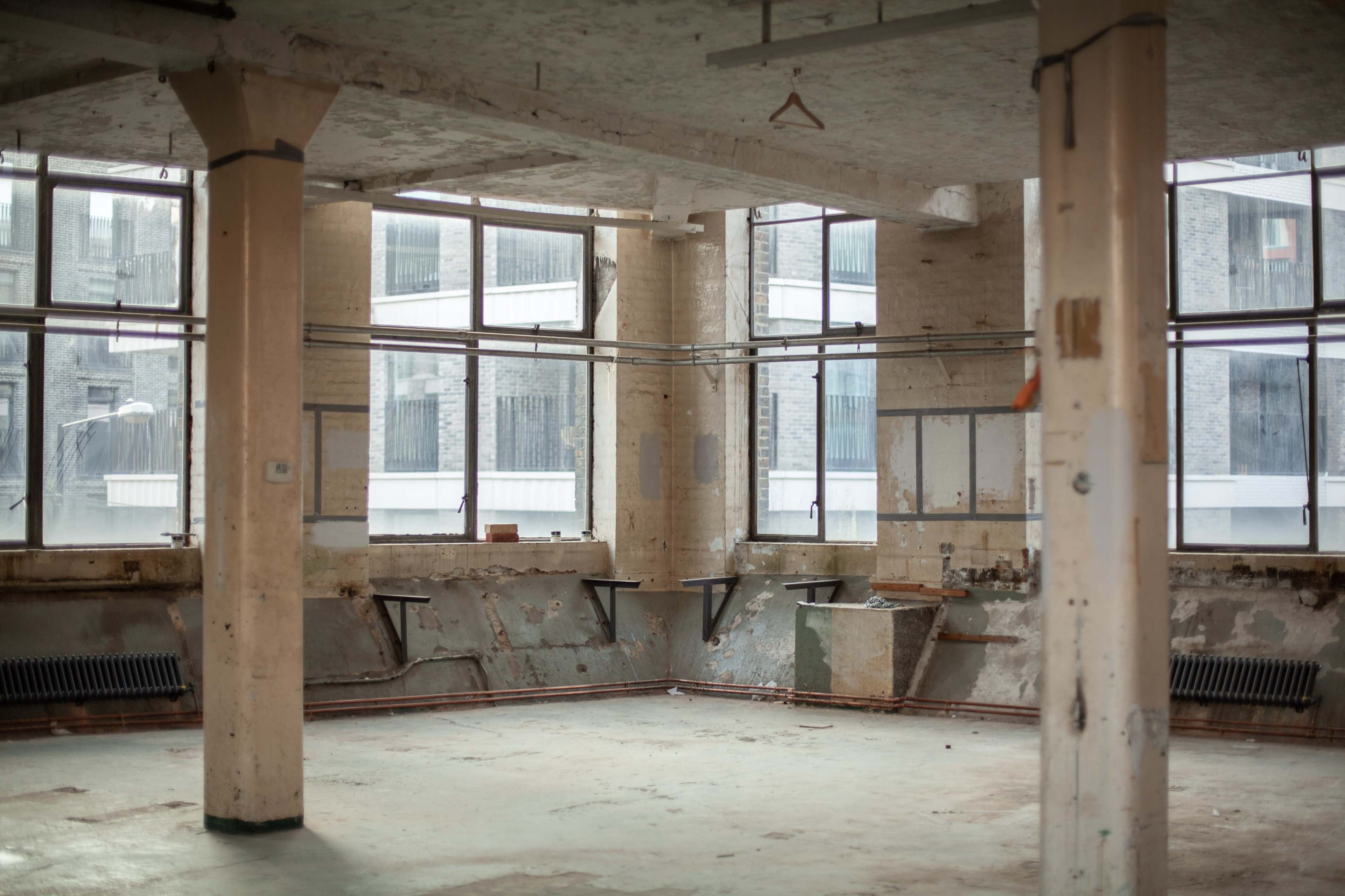 The image shows an empty, industrial space with bare walls, large windows, and exposed pipes.