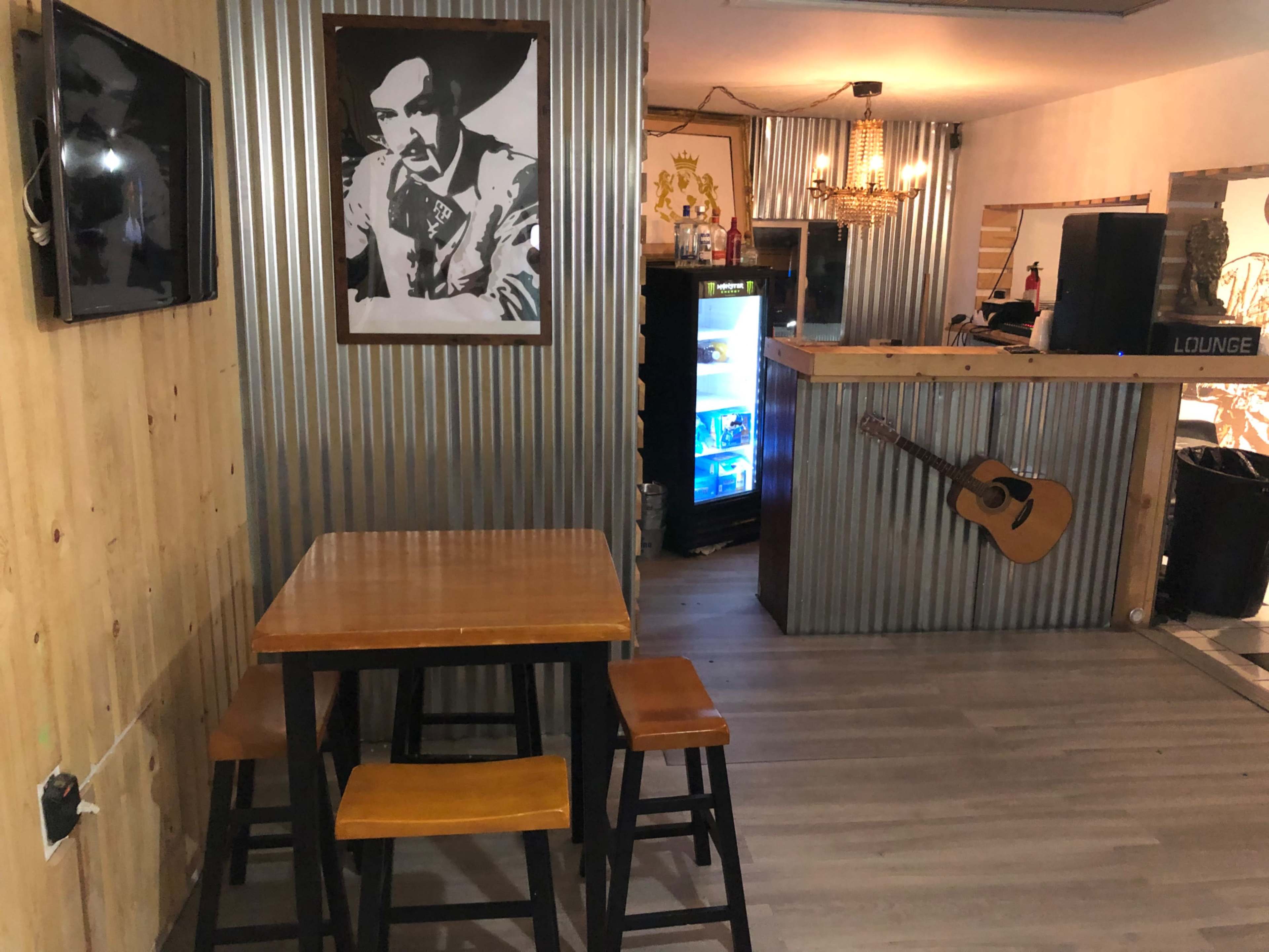 A casual lounge area with wooden furniture, a corrugated metal bar, and a framed portrait on the wall.