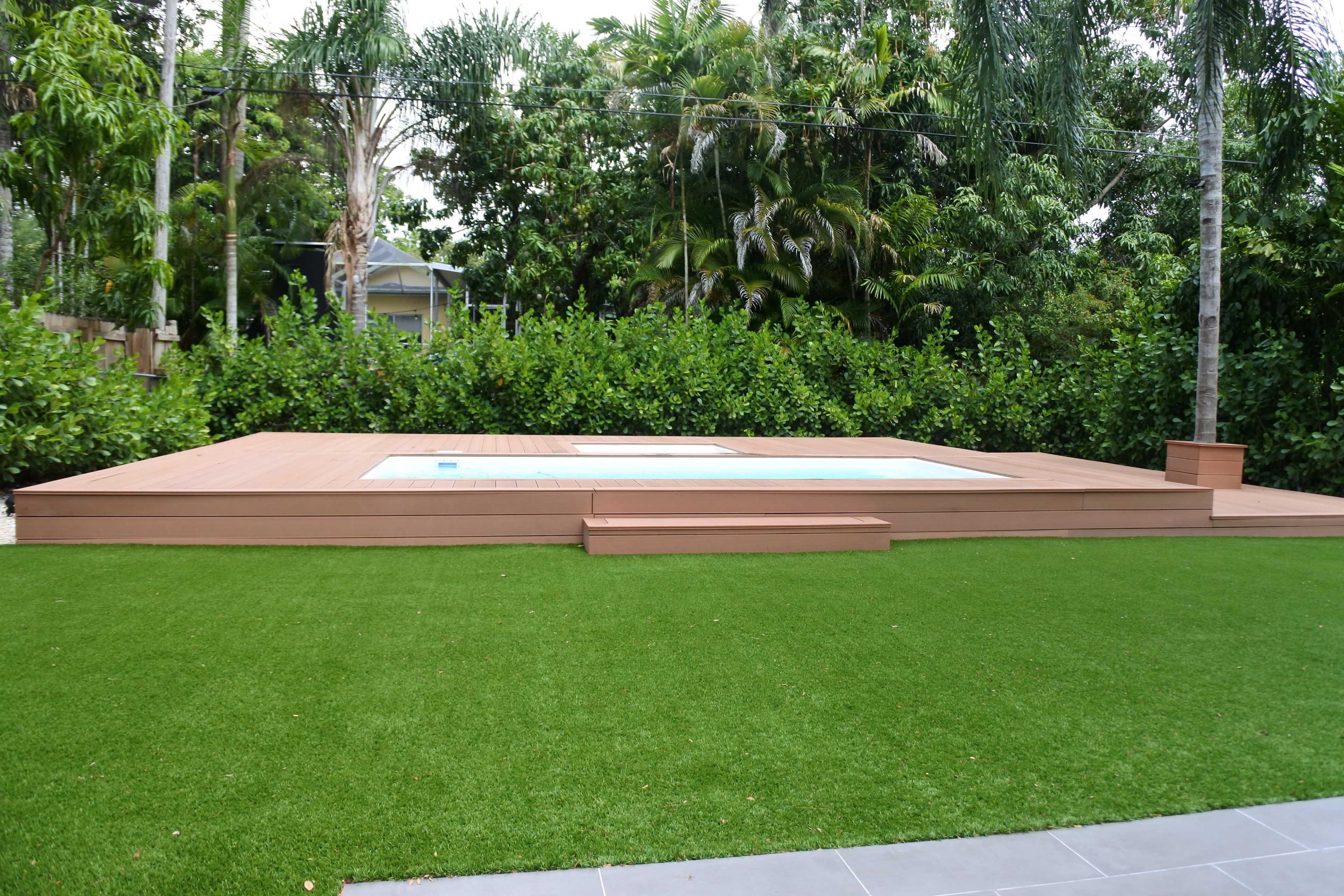 A wooden deck with a sunken pool surrounded by artificial grass and tropical foliage.