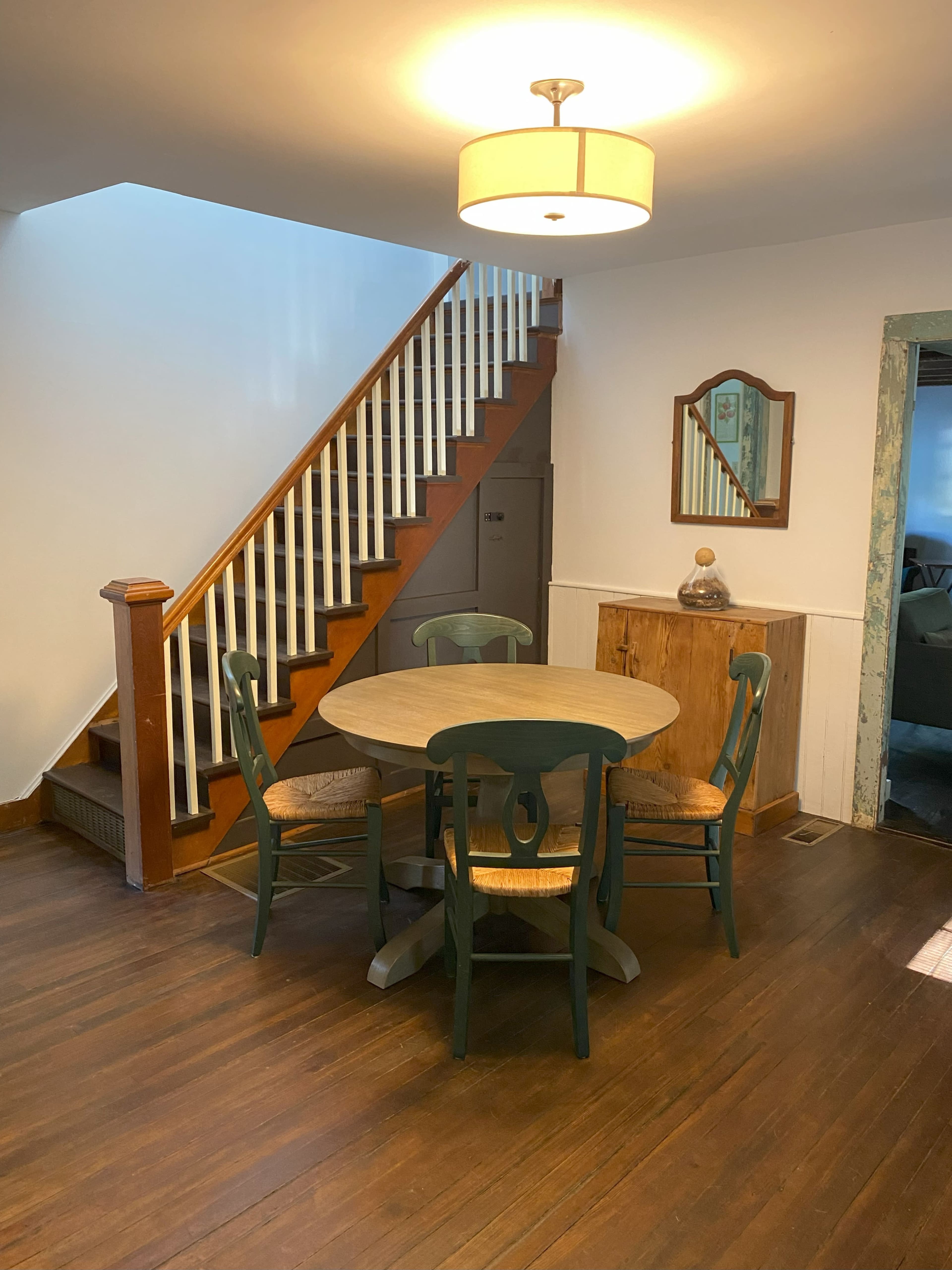 A round dining table with four chairs is set against a wall near a staircase in a well-lit room with wooden flooring.