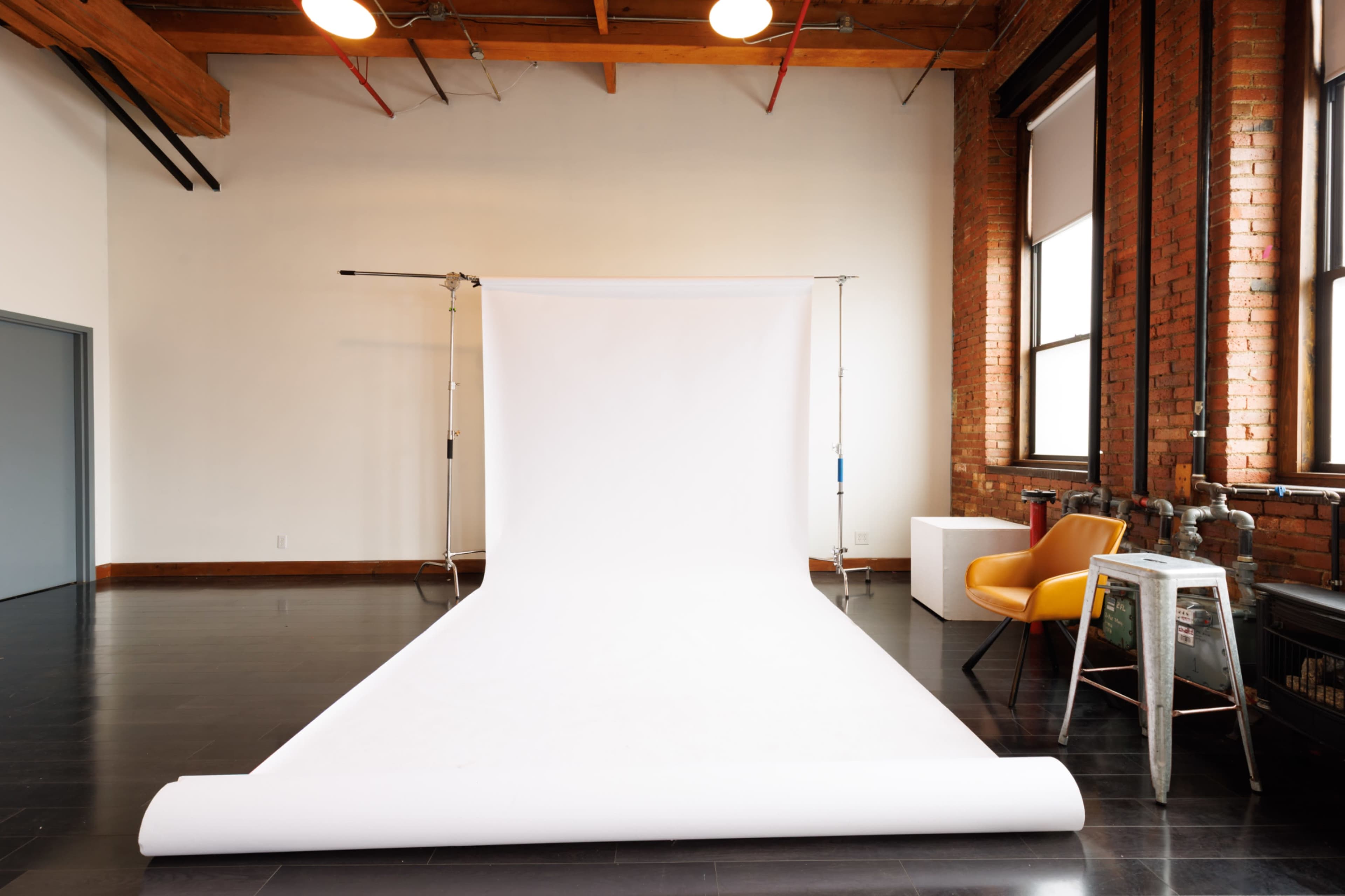 A photography studio features a large white backdrop roll, an orange chair, and exposed brick walls.