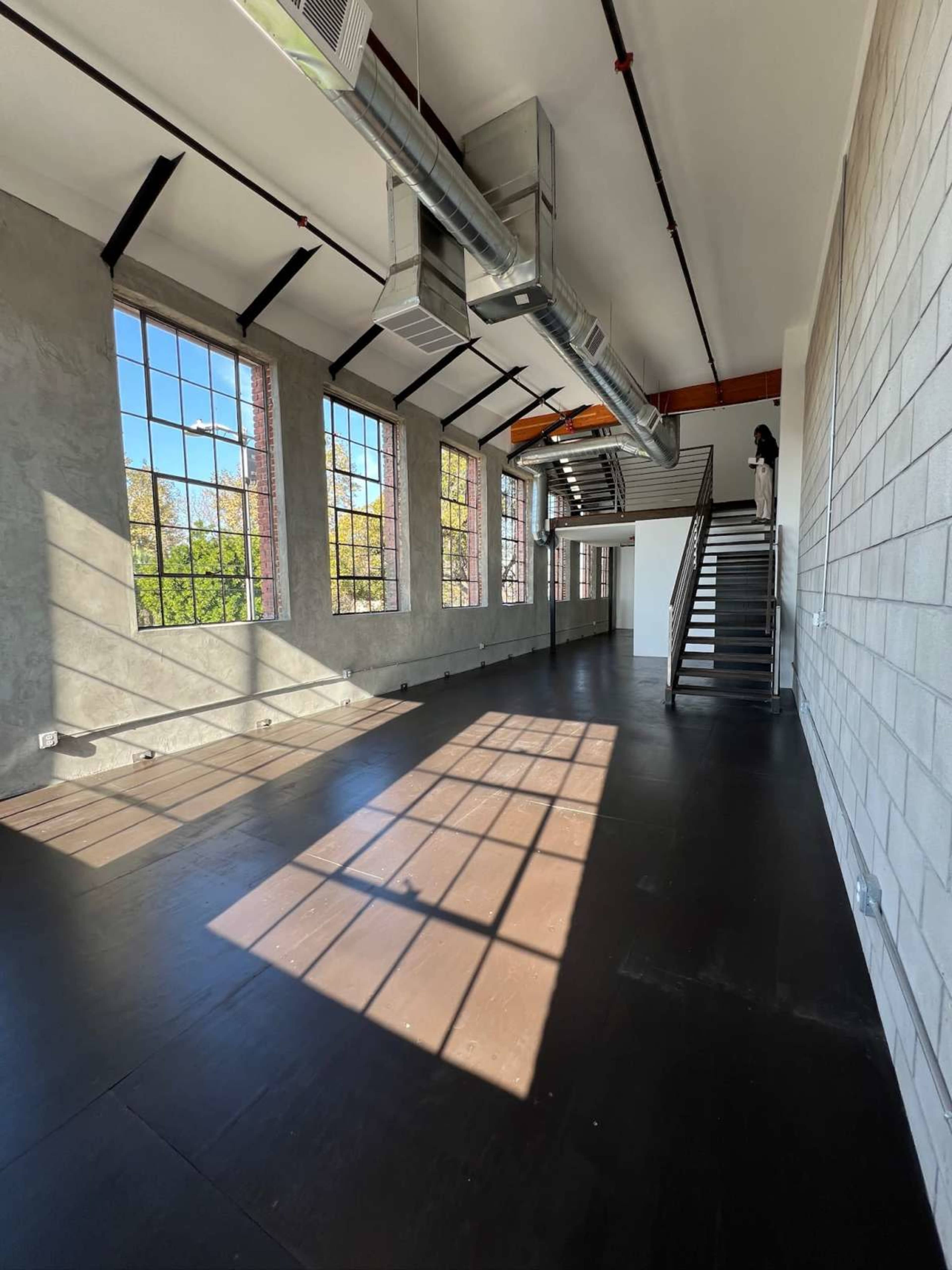 The image shows a spacious industrial-style room with large windows, black flooring, and metal ductwork on the ceiling.