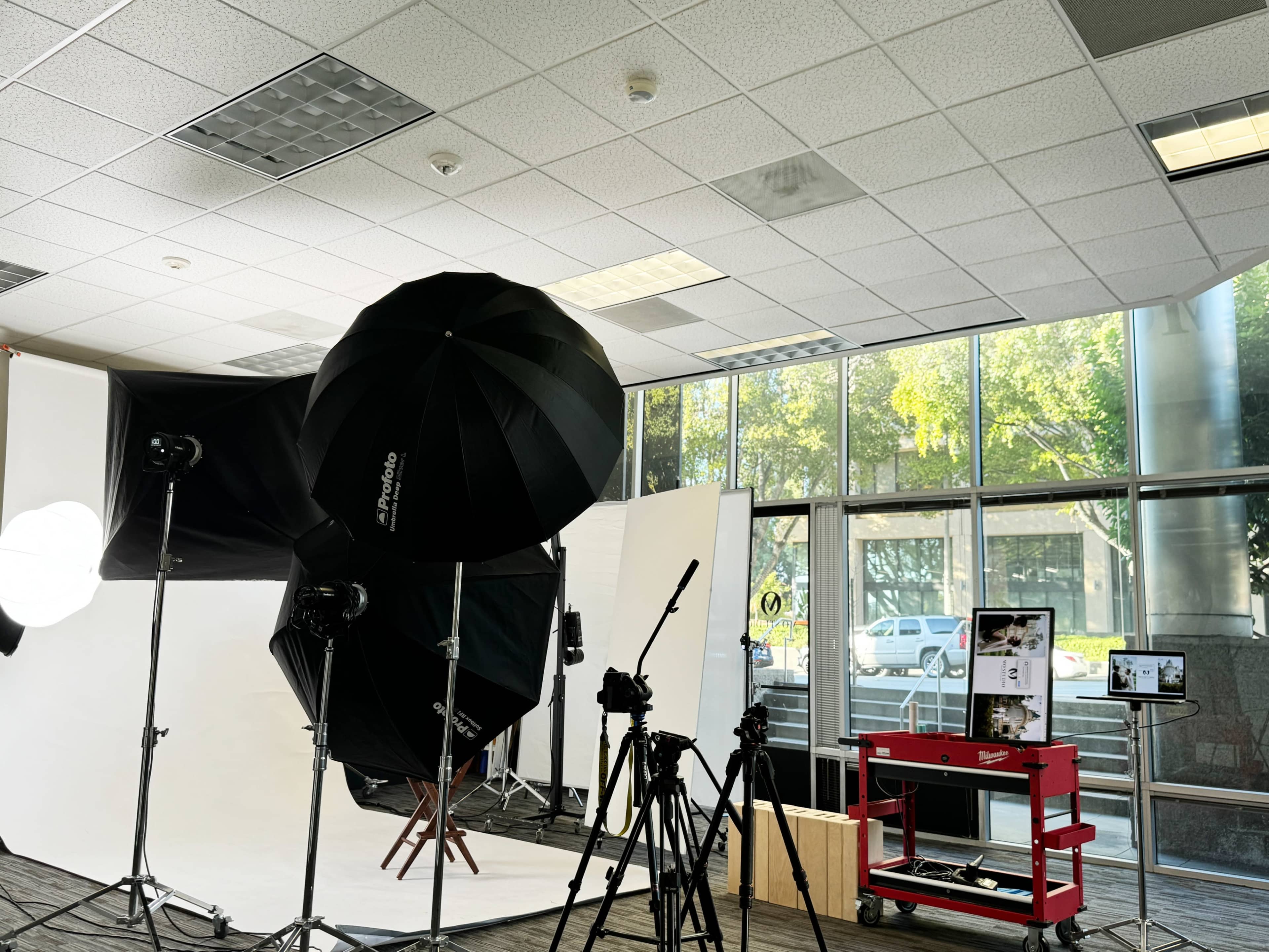The image shows a photography studio with various lighting equipment, backdrops, and cameras set up next to large windows.