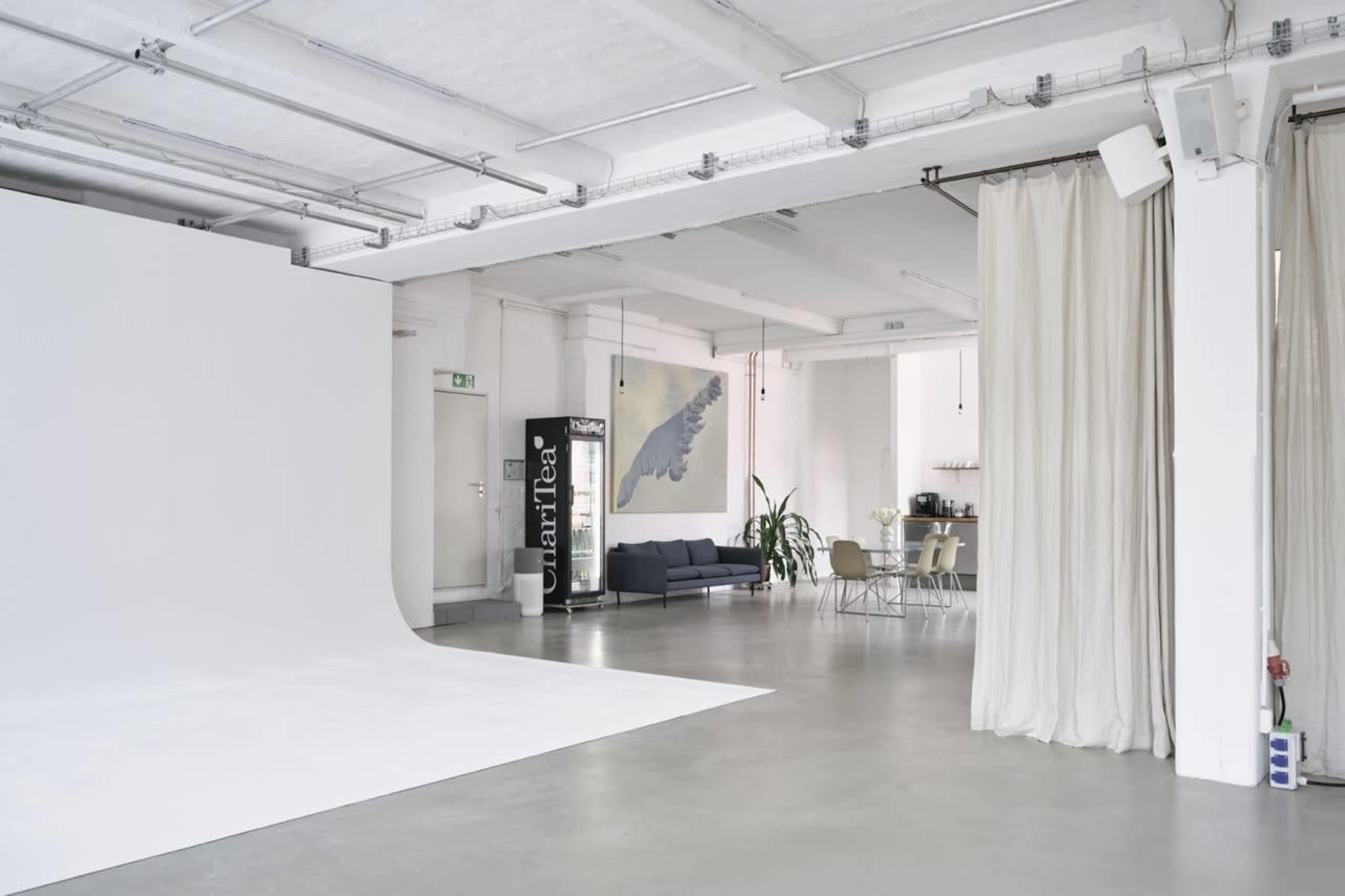 The image shows a spacious, minimalist studio with a white backdrop, a dark sofa, a dining area, and large curtains.