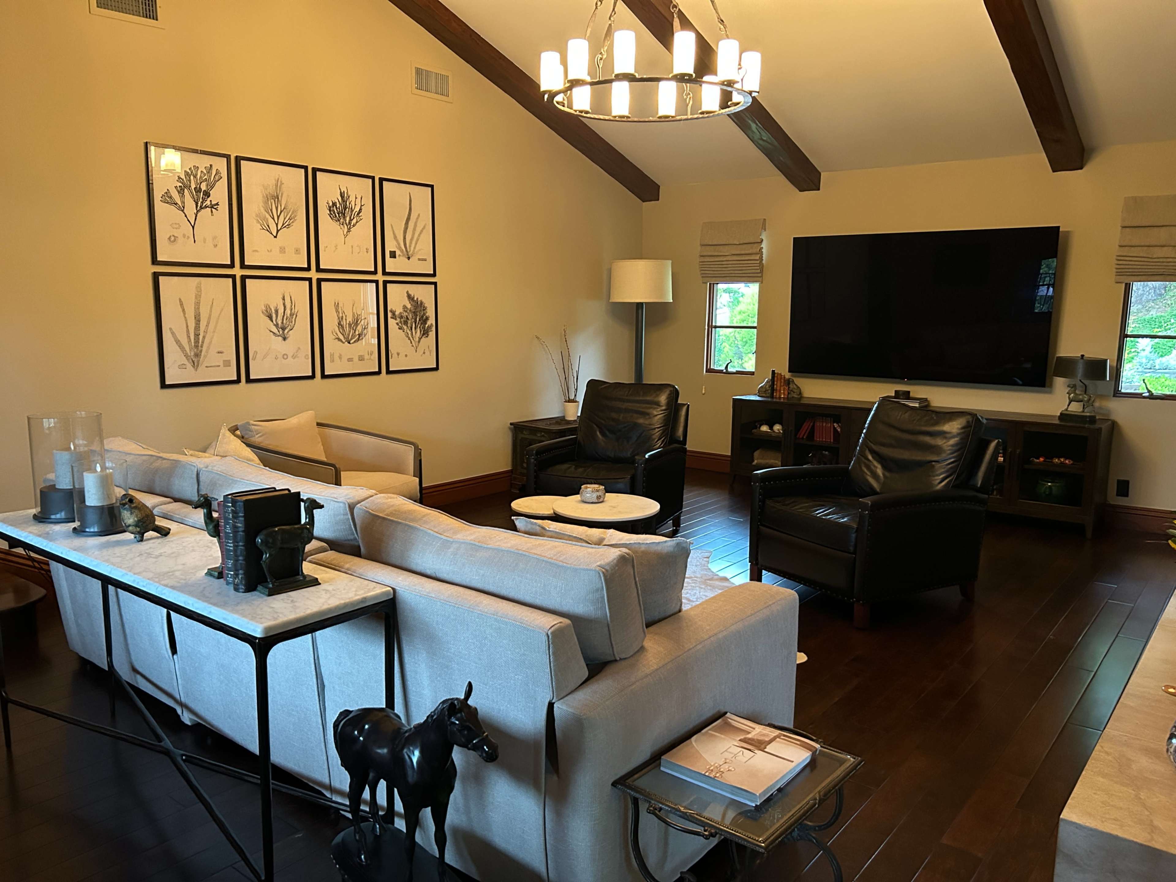 The living room features a beige sectional sofa, two black leather armchairs, a coffee table, a large television mounted on the wall, and framed botanical prints.