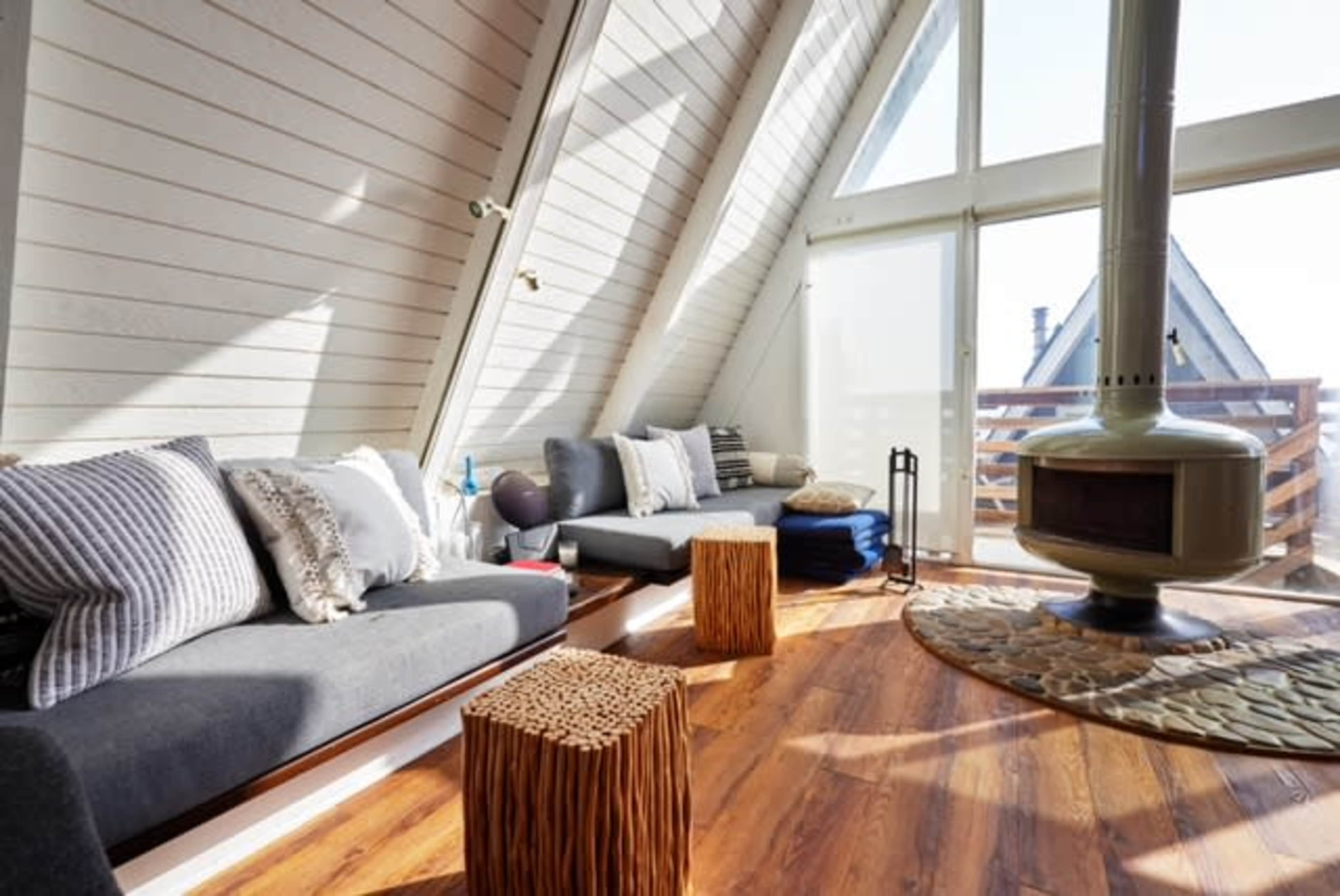 The bright living room features slanted wooden walls, two sofas with pillows, and a central stone fireplace, all complemented by natural light streaming through large windows.