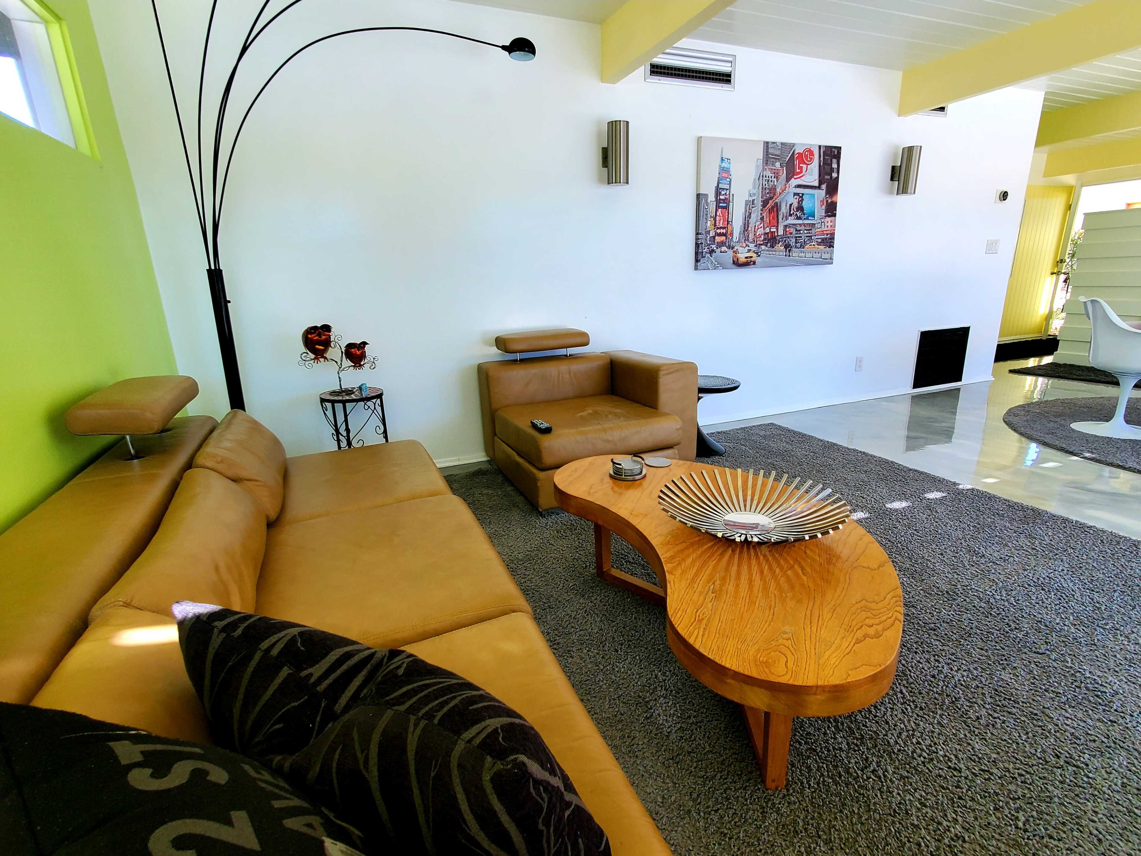 The image shows a modern living room featuring a tan leather sectional sofa, a wooden coffee table, and a light green accent wall, with a large piece of art depicting a cityscape on the wall.