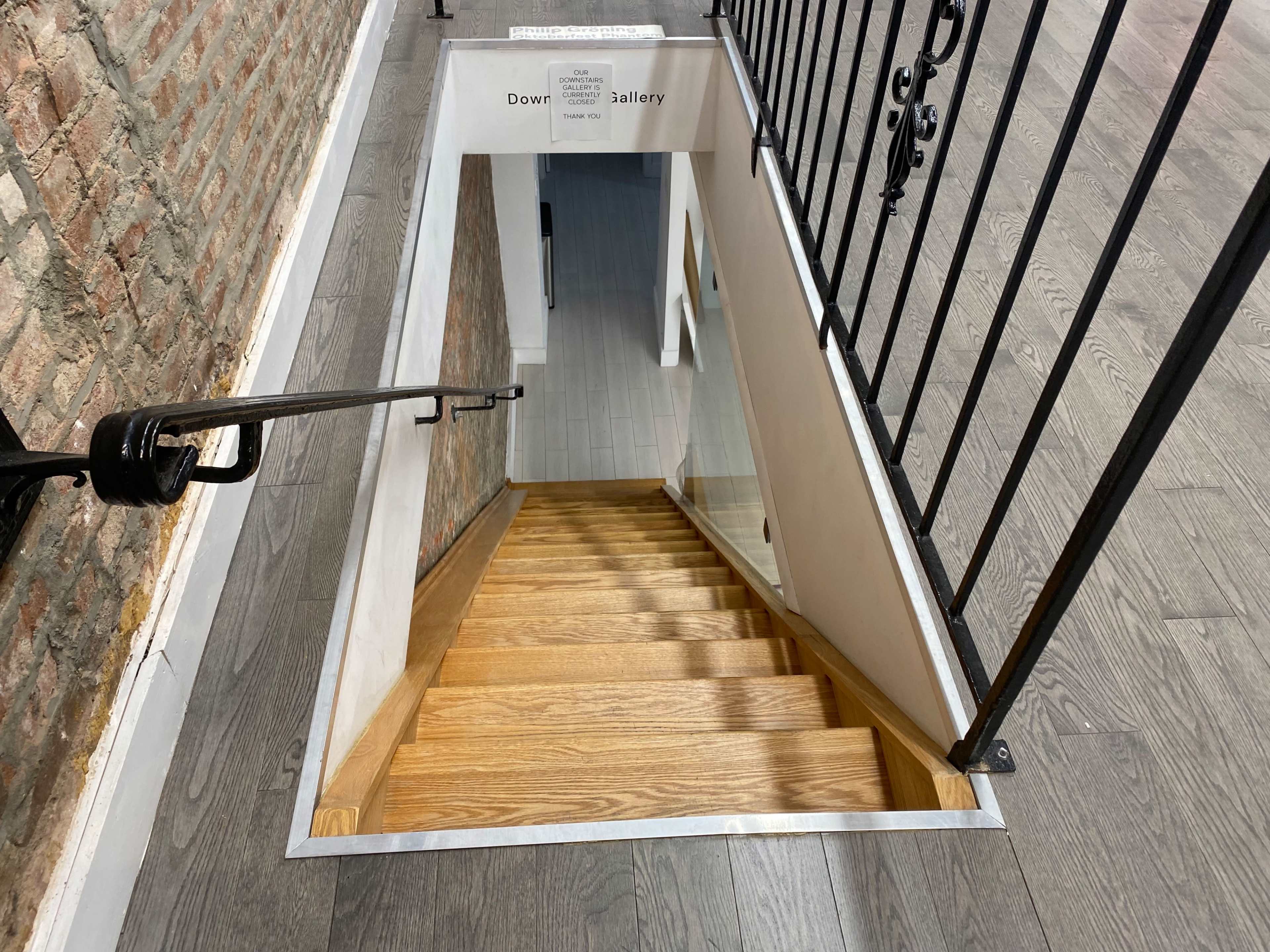 A staircase with wooden steps descends into a well-lit space beneath a sign indicating "Down to Gallery."