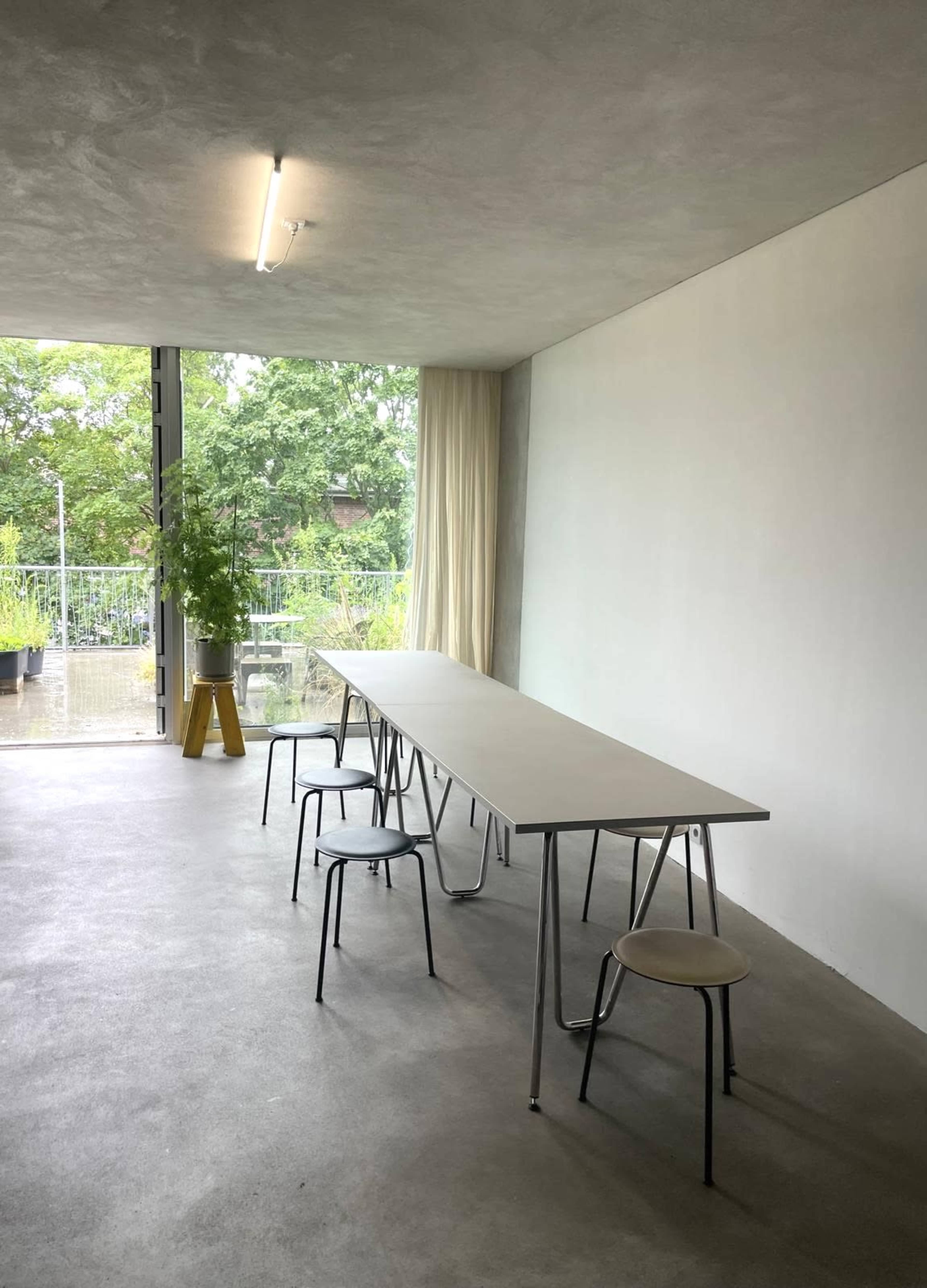 A long, rectangular table with several chairs is set up in a bright room featuring large windows that overlook greenery outside.
