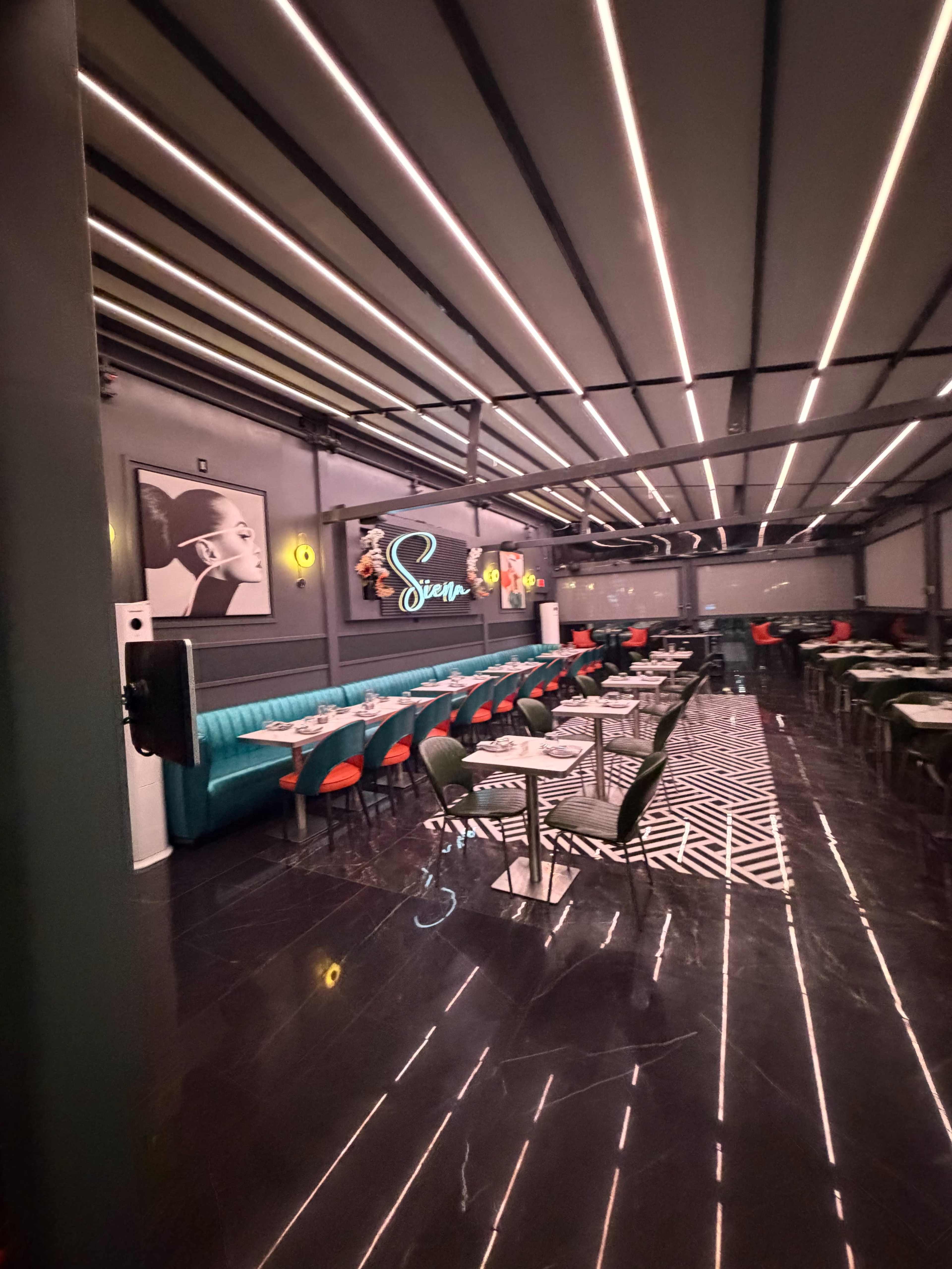 The image shows a modern restaurant interior featuring a mix of green and red chairs, sleek tables, and decorative lighting along the ceiling.