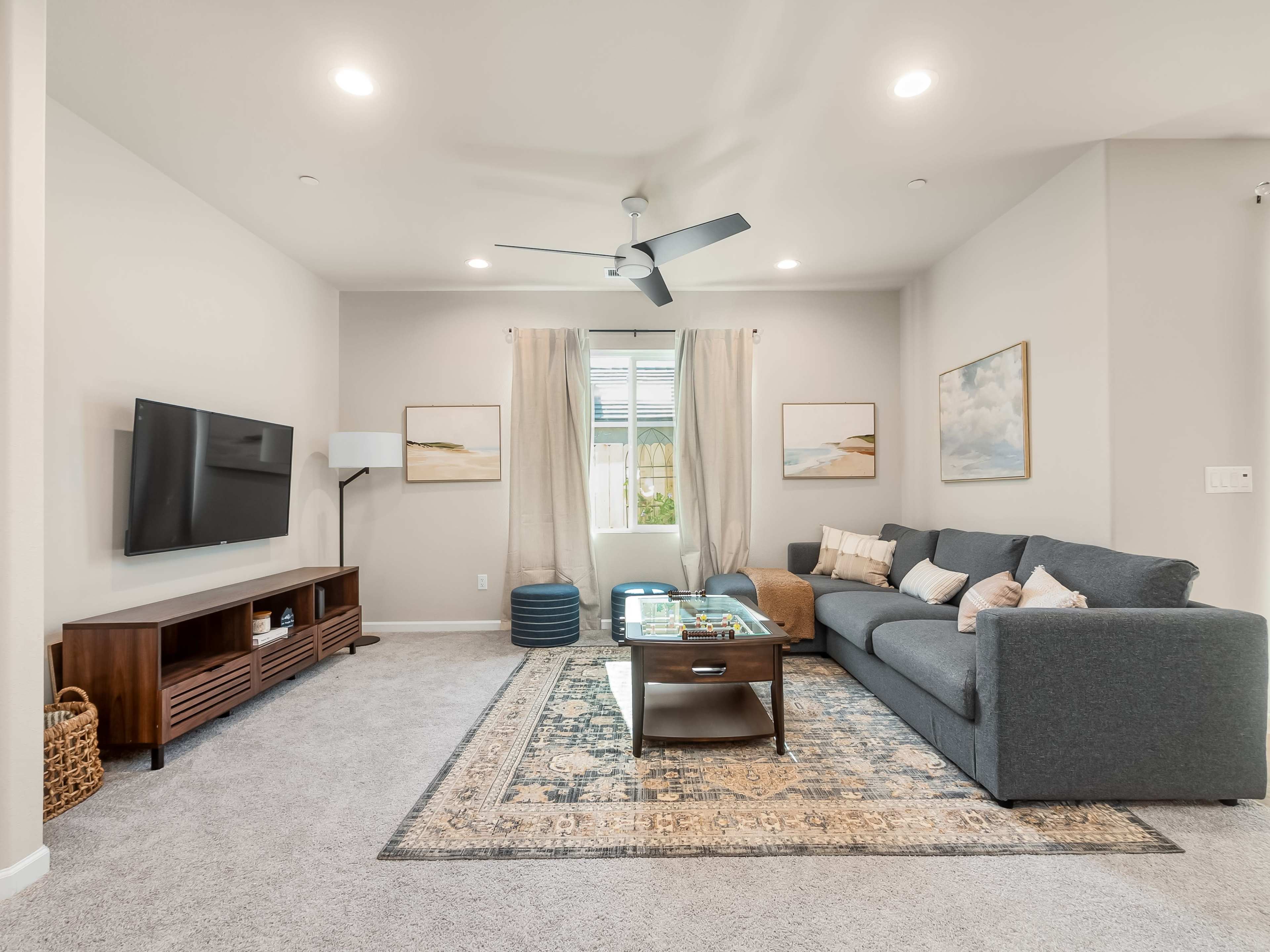 A modern living room features a gray sofa, a wooden TV stand, and a coffee table on a patterned rug, with large windows allowing natural light.