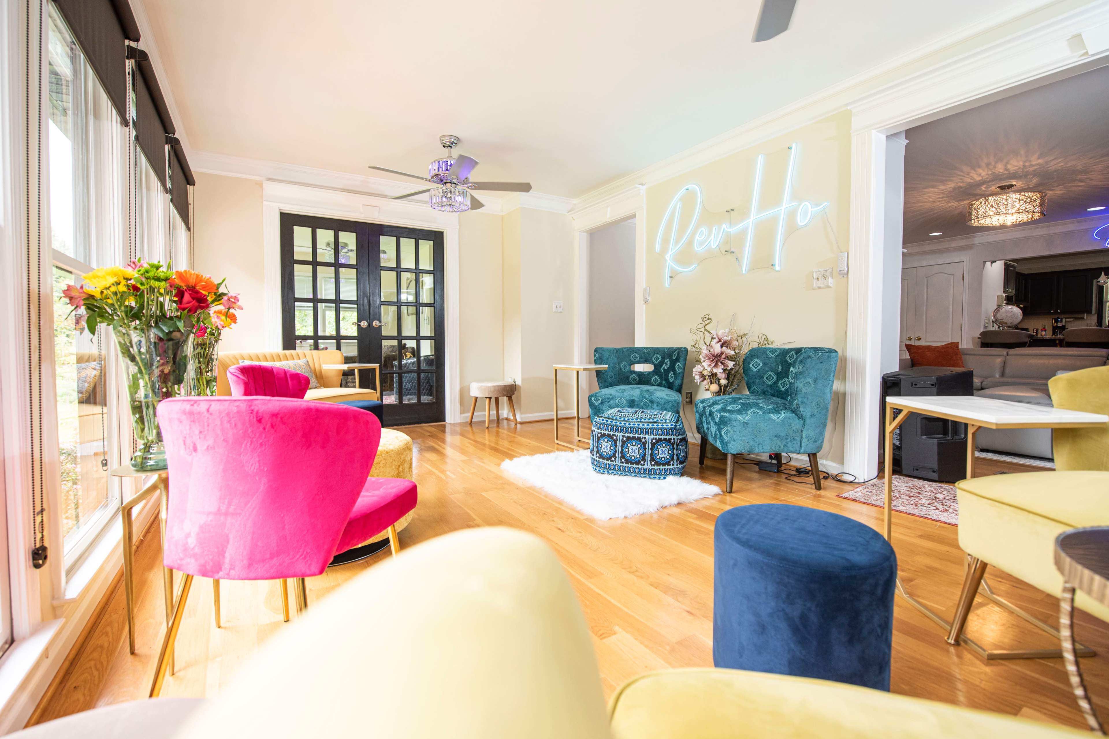 A bright seating area features colorful chairs, a decorative table, and a central rug, with a neon sign on the wall.
