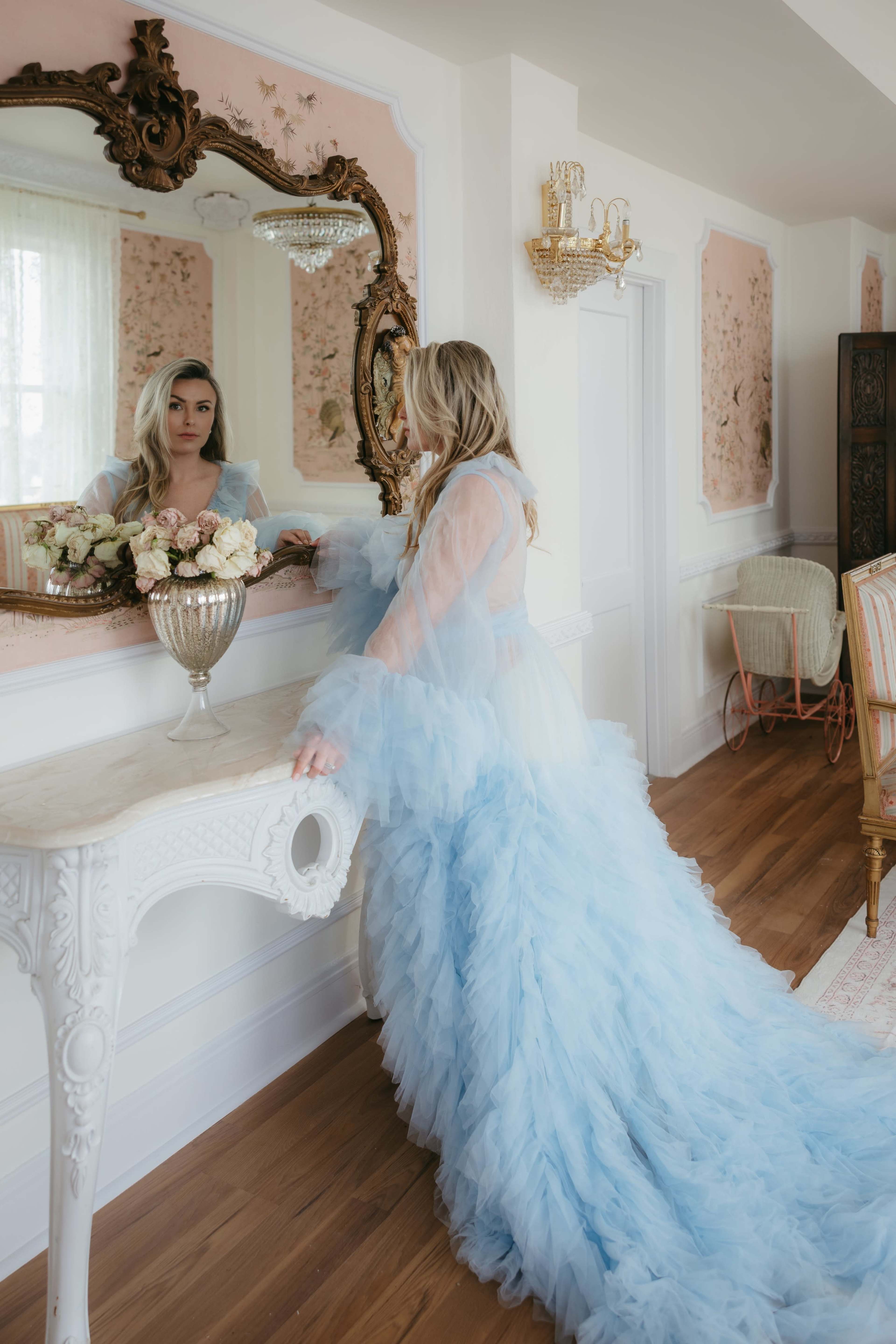 A woman in a long, ruffled blue gown stands in front of an ornate mirror, adjusting her hair in a softly lit room with floral wallpaper and elegant furnishings.