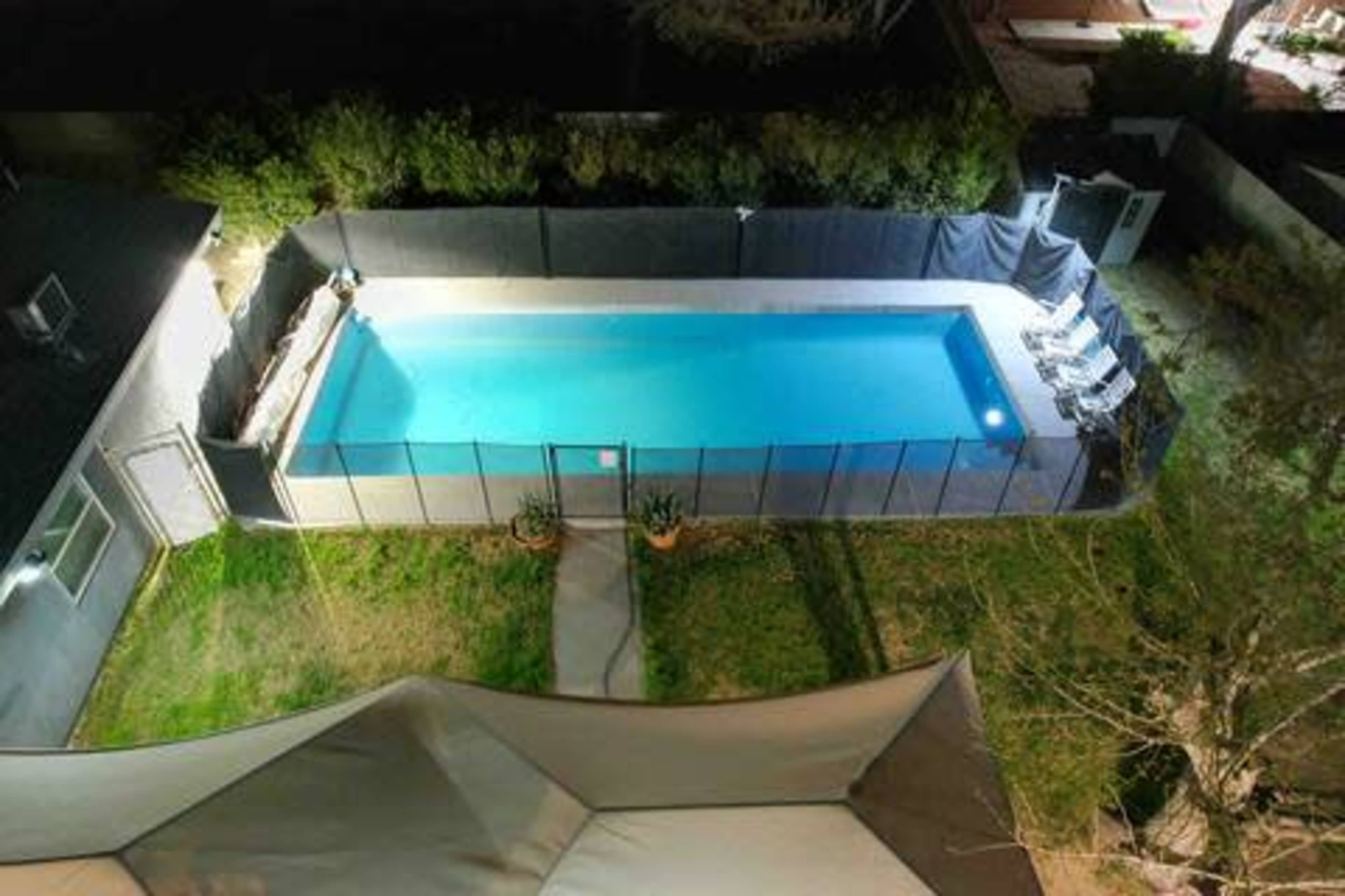 An aerial view shows a rectangular swimming pool surrounded by a fence and lounge chairs, with grass and a pathway leading to a house.