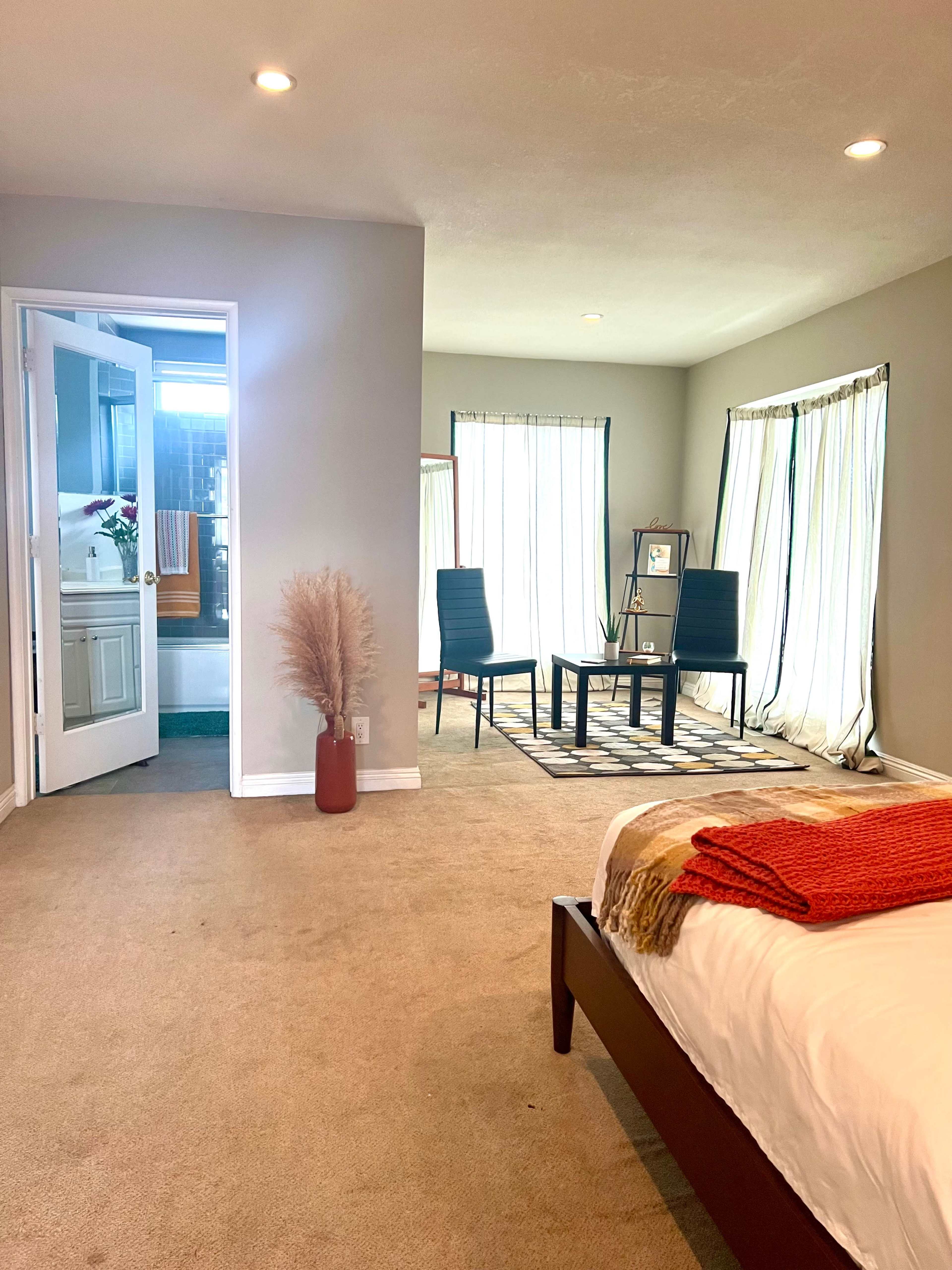 A spacious bedroom features a bed with an orange throw, a small sitting area with two chairs and a table, and doors leading to a bathroom.