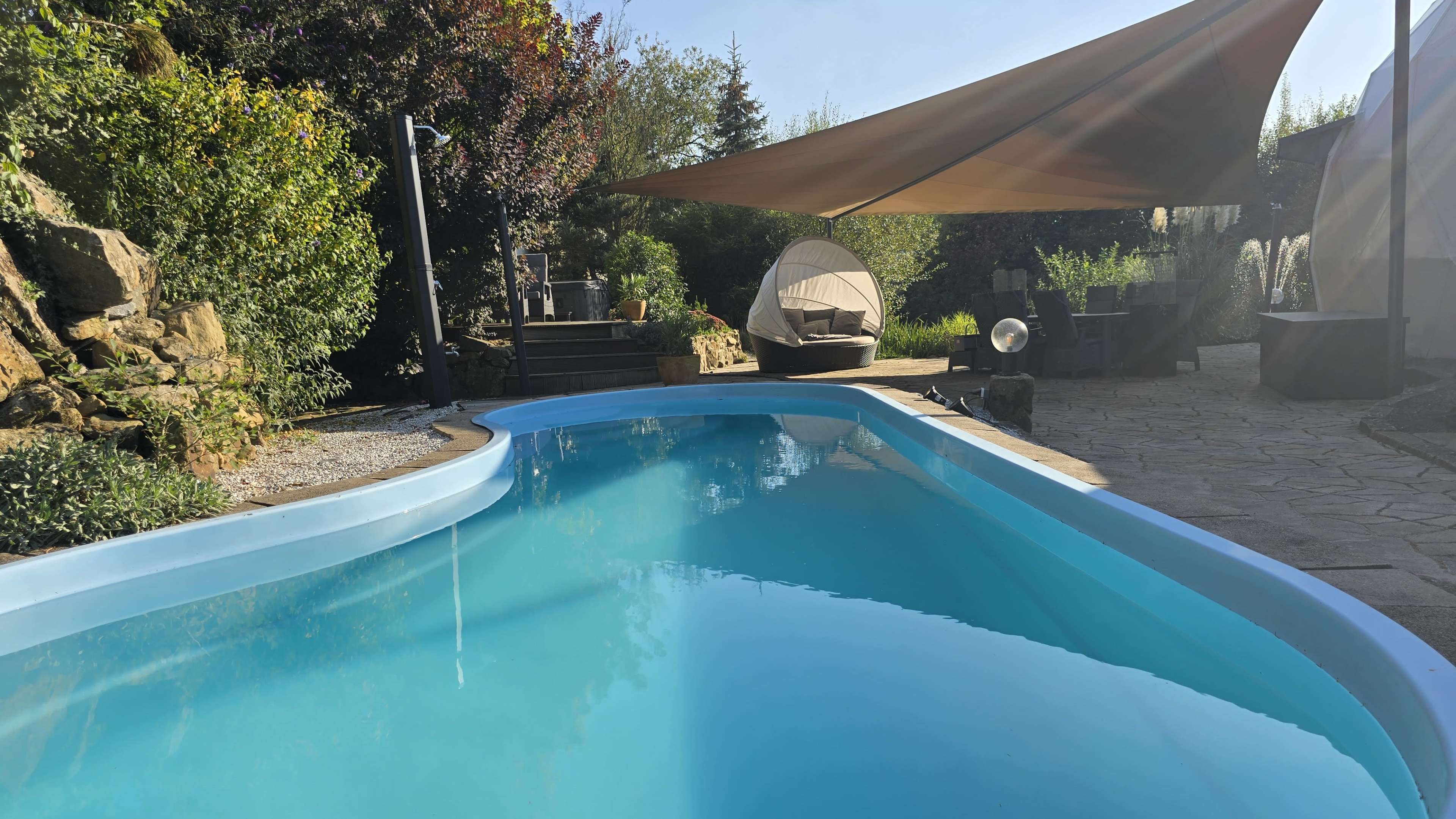 A clear blue swimming pool is surrounded by stone pathways and greenery, with a shaded seating area in the background.