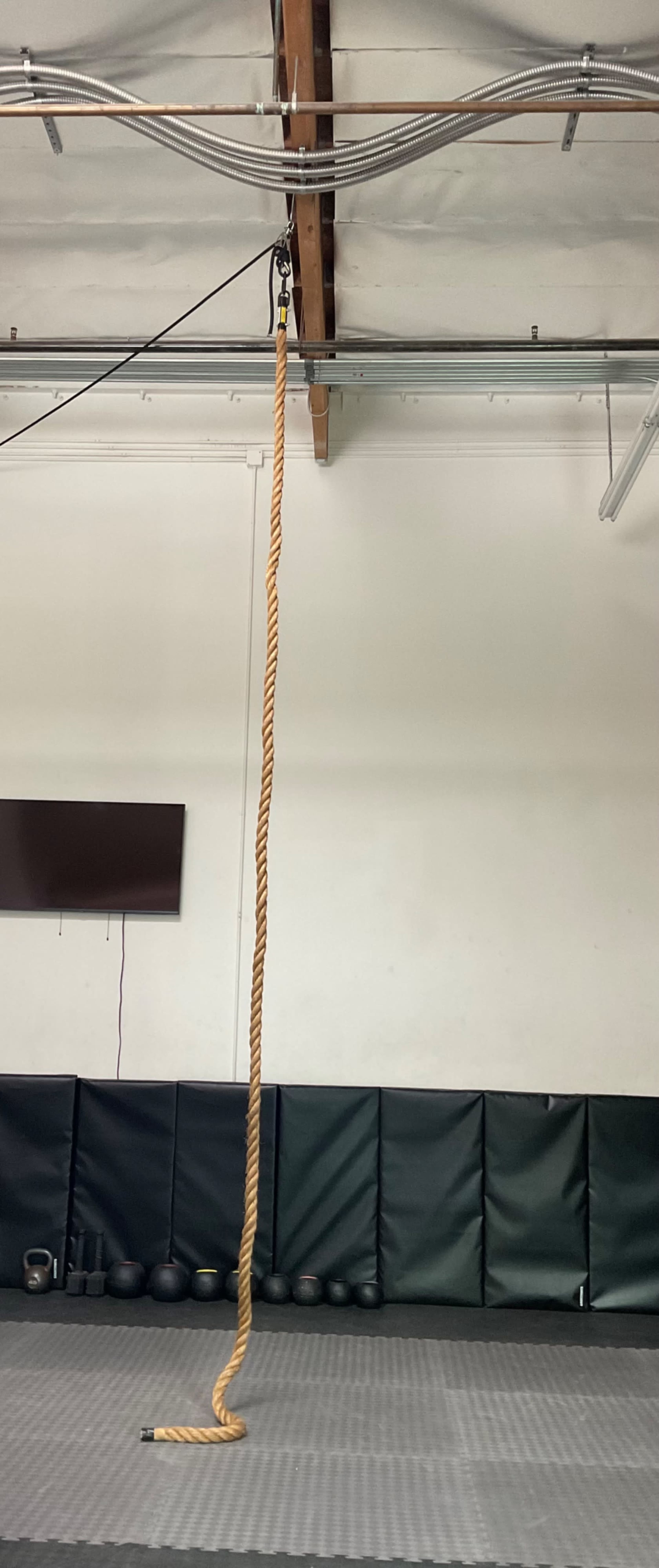 A thick climbing rope hangs from a ceiling fixture in a gym with black mats and exercise equipment visible in the background.