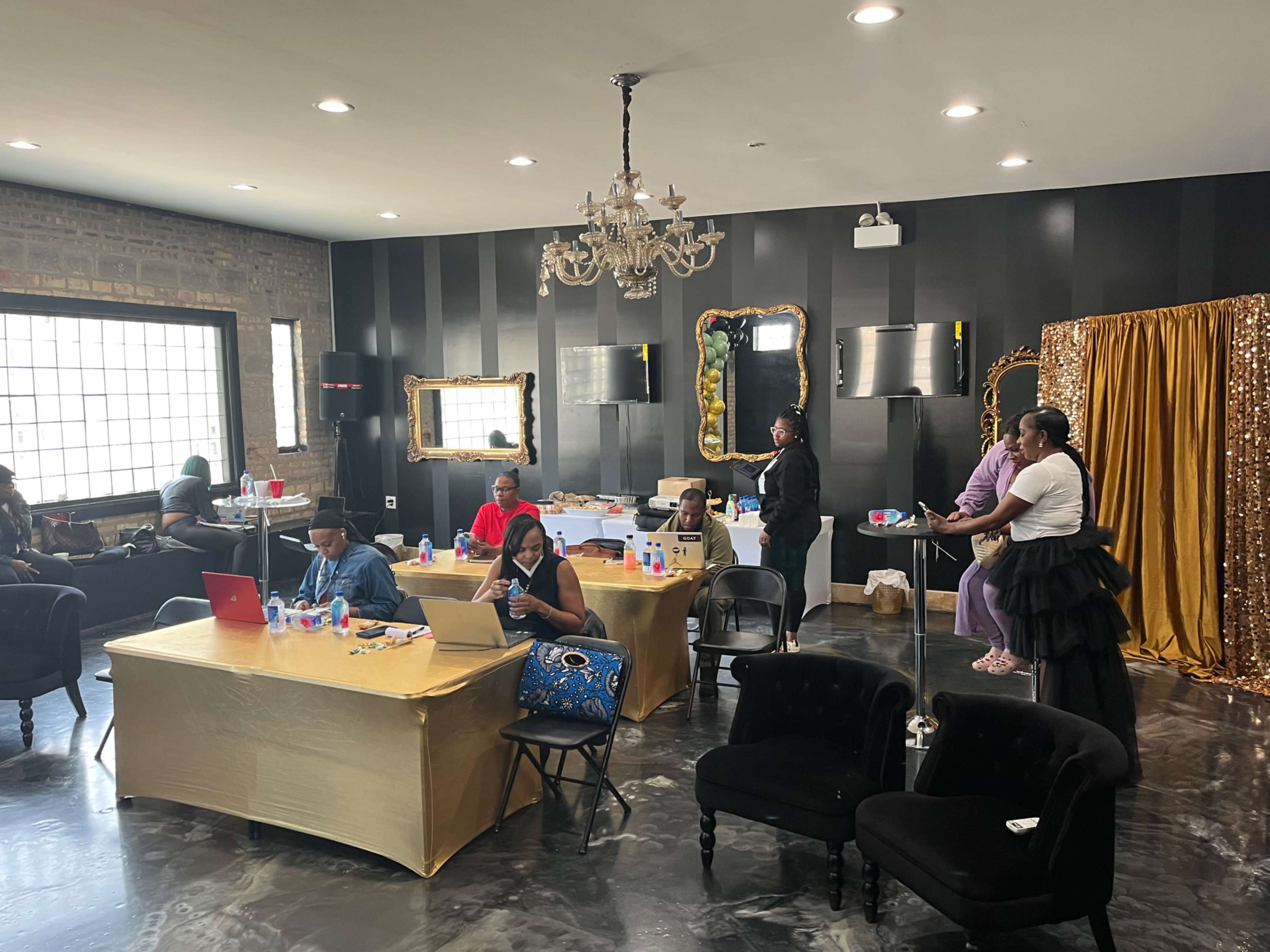 A group of people is gathered in a spacious room with black walls and decorative mirrors, working at tables and interacting near a gold curtain backdrop.
