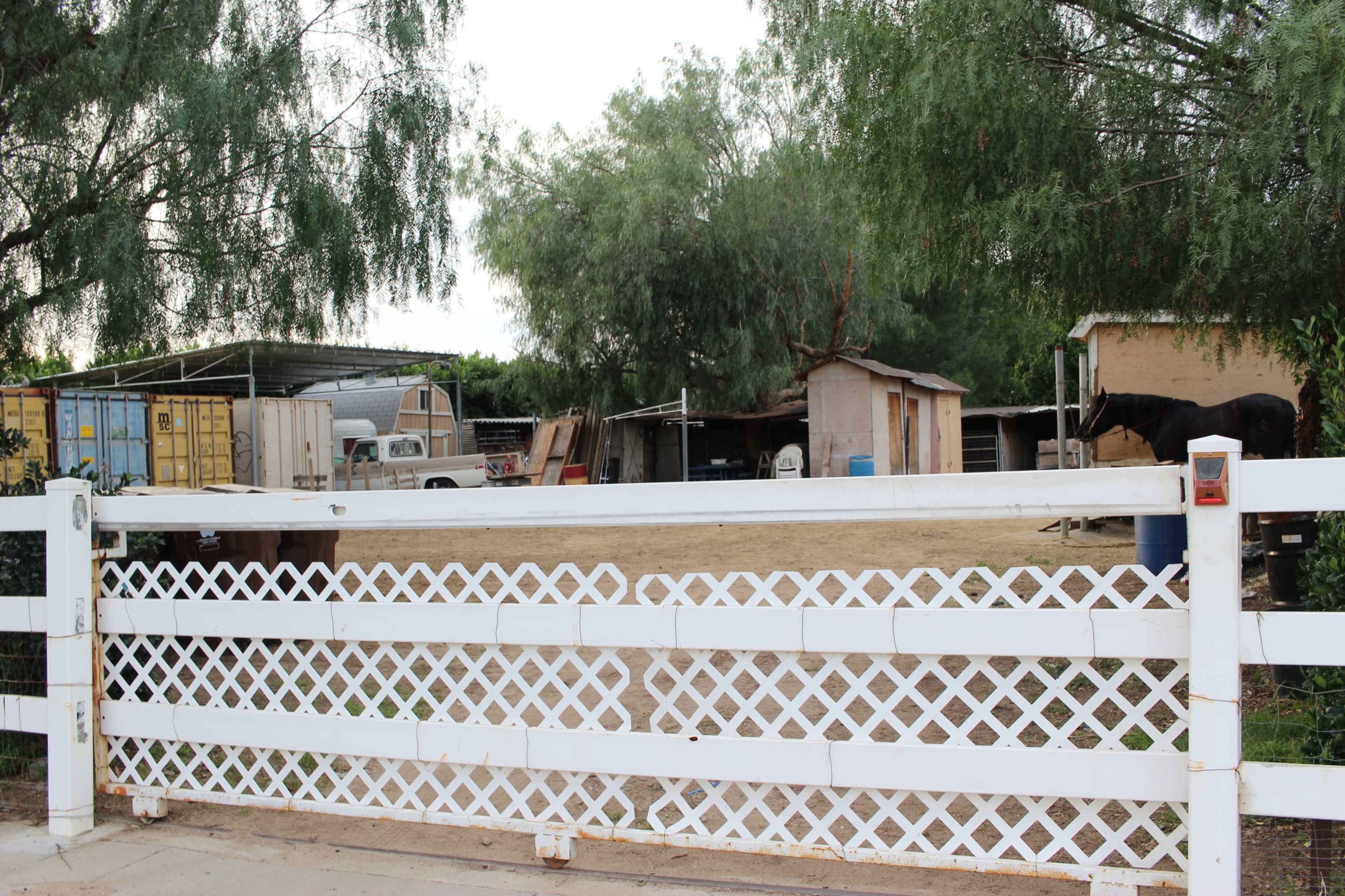 The image shows a white picket gate leading to a farm area with structures, vehicles, and a horse in the background.