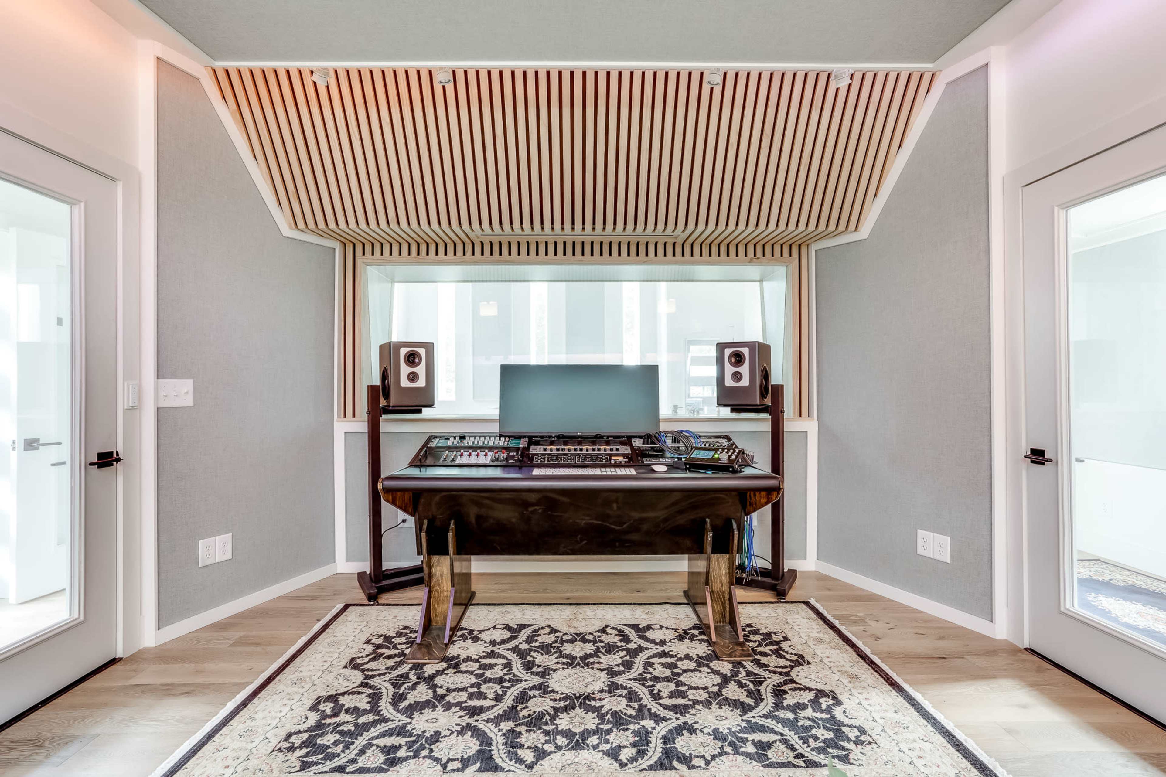 A modern music studio with a wooden mixing desk, speakers on either side, and large windows allowing natural light.