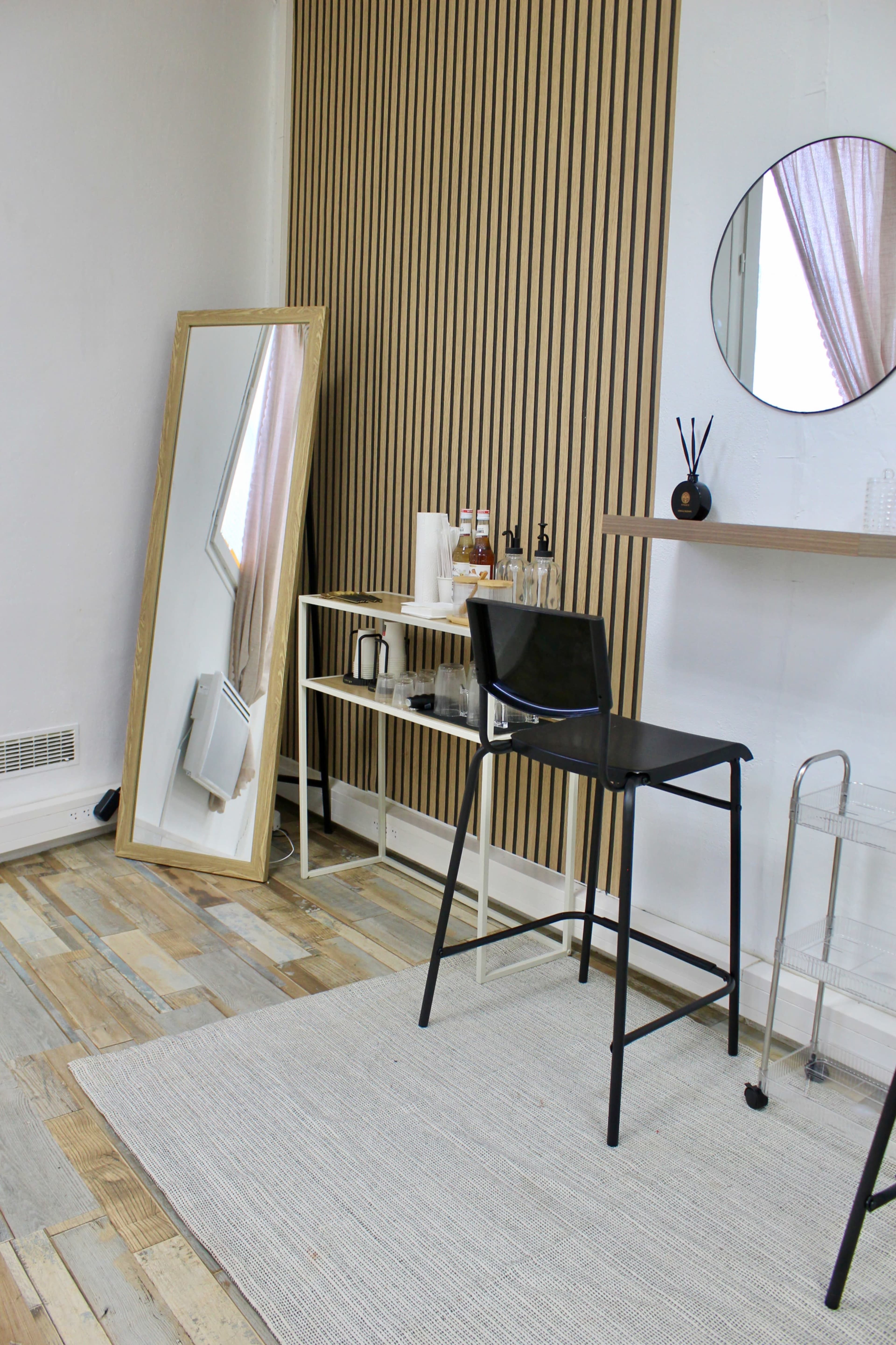 The image shows a minimalist interior featuring a tall black stool, a large mirror against a wooden slatted wall, and a small shelf with various items.