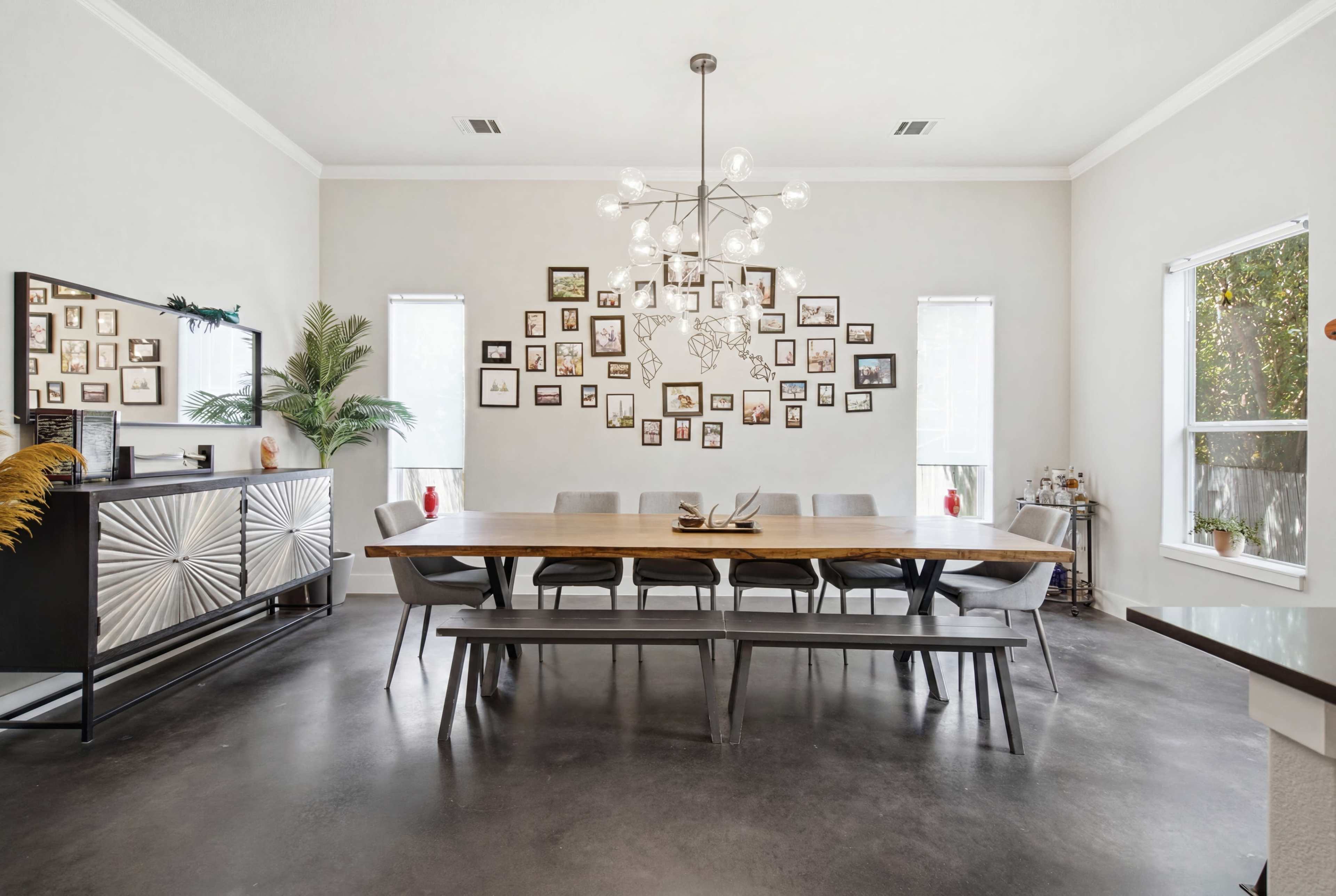 A spacious dining room features a long wooden table surrounded by gray chairs, with a gallery wall of framed photos and a modern chandelier above.